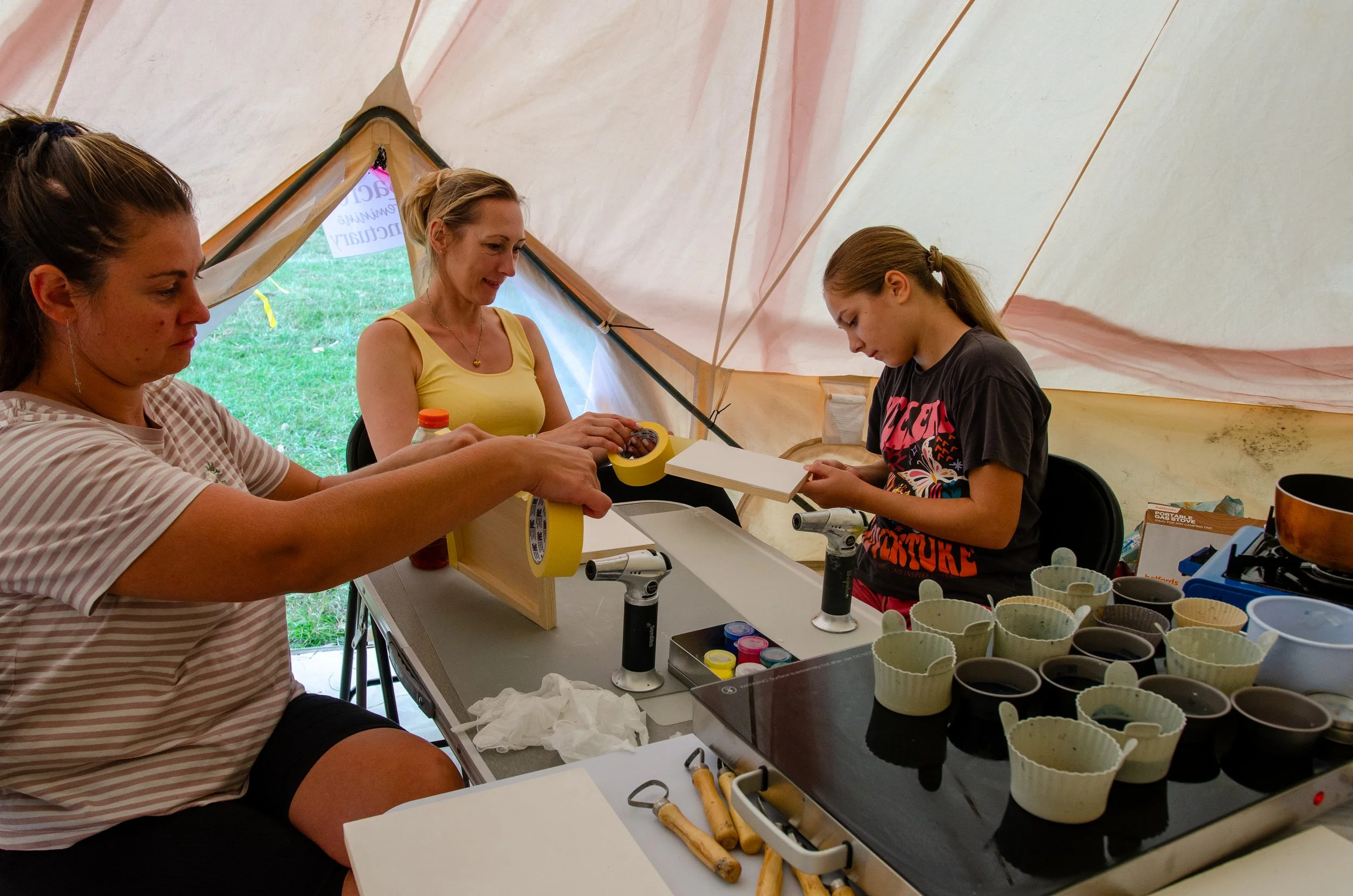 Three women and a girl working on a craft project inside a tent, surrounded by bowls and tools.