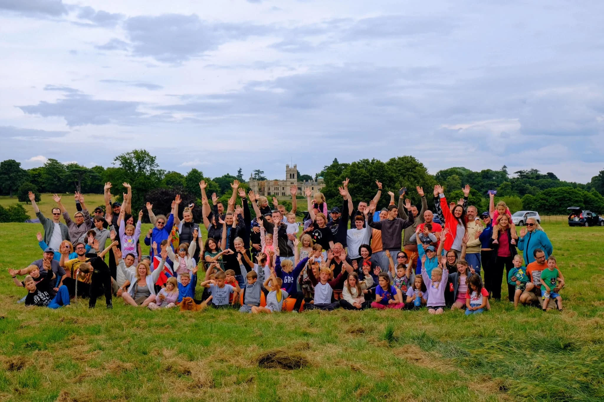 A large group of people, including children and adults, gathered outdoors on a grassy field, with many raising their hands in the air. There is a castle or large historic building in the background, surrounded by trees and a cloudy sky.