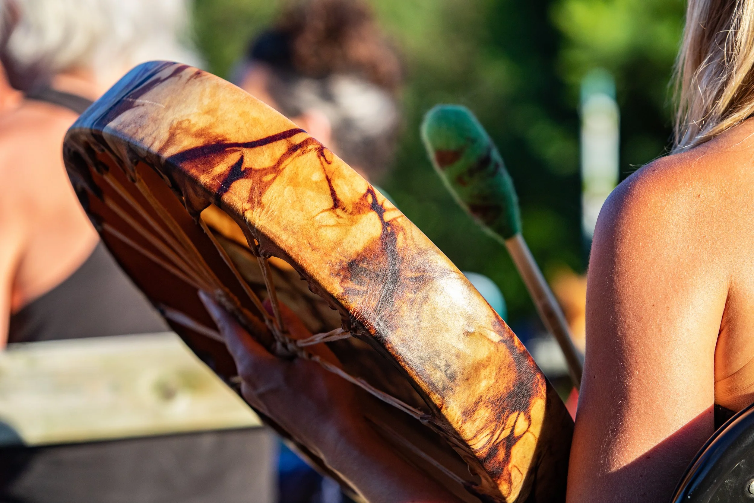 Close-up of a woman holding a drum at an outdoor event, with other people and leafy background in focus.