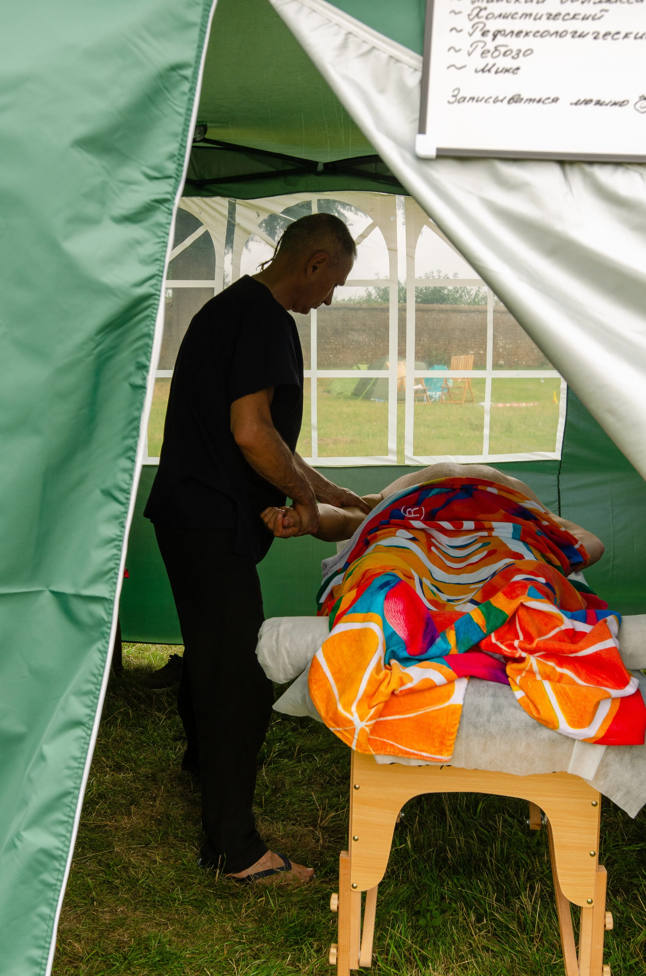 A person receiving a massage outdoors inside a green tent, with a whiteboard displaying handwritten notes in the background.