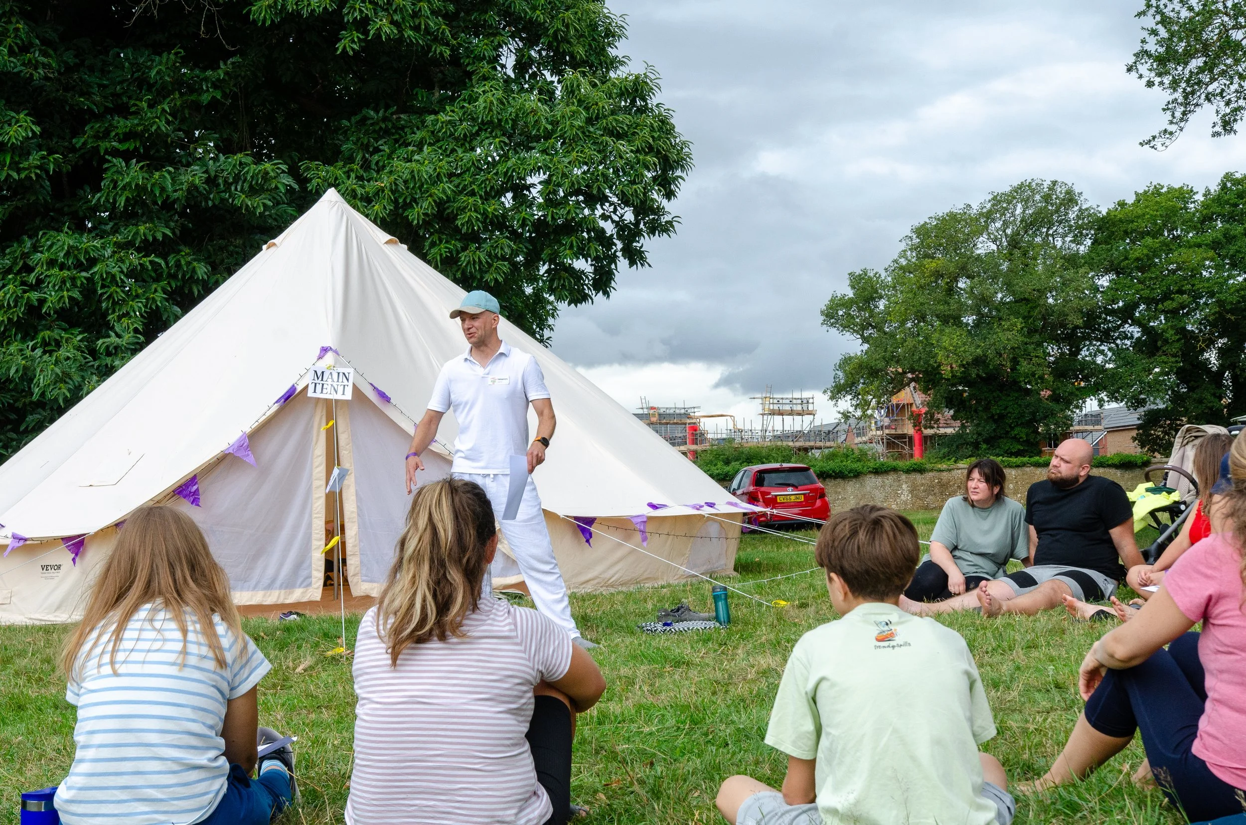A group of children and adults sitting on the grass around a man in white clothes giving a presentation outdoors in front of a large white tent, under cloudy skies.