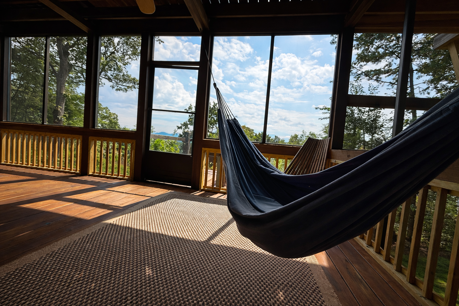 A screened-in porch with wooden flooring and railing, a hammock hanging, and a view of trees and blue sky with clouds outside.