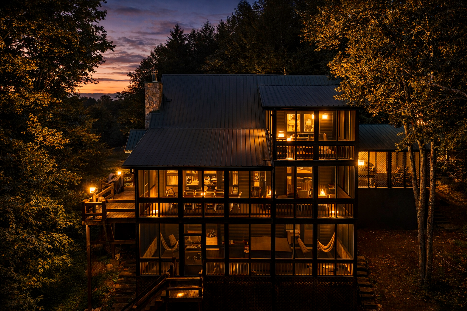 A multi-story house with large glass windows and balconies, illuminated from within, surrounded by trees at dusk.