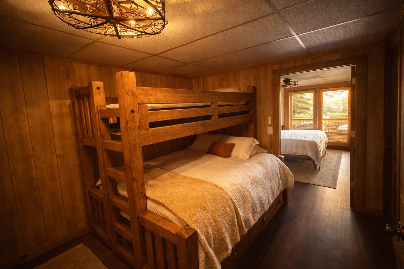A cozy bedroom with wooden paneling walls, featuring a bunk bed above a double bed. Another bedroom with large windows and views is visible through an open doorway.