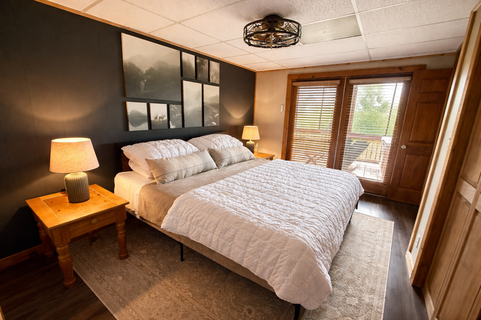 A cozy bedroom with a large bed, two bedside tables with lamps, wooden floors, a dark wood accent wall with artwork, and balcony doors with wooden blinds letting in natural light.