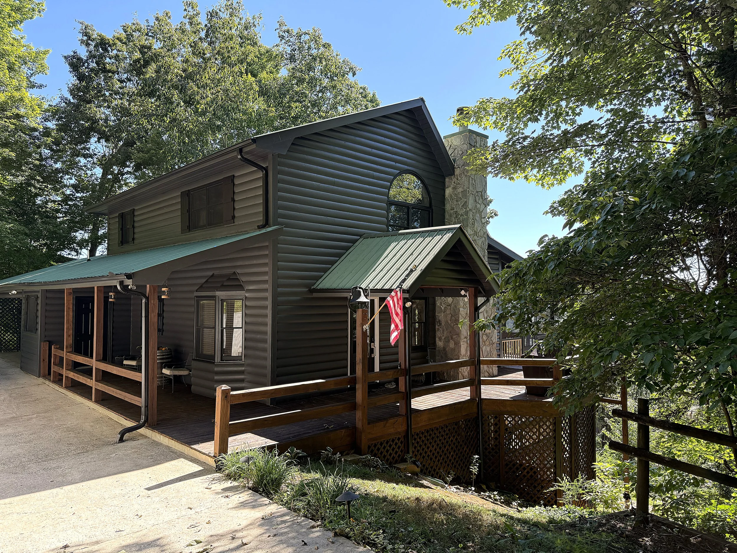 A multi-level house with dark siding and a green metal roof, surrounded by trees, with a wooden deck and railing, under a clear blue sky.