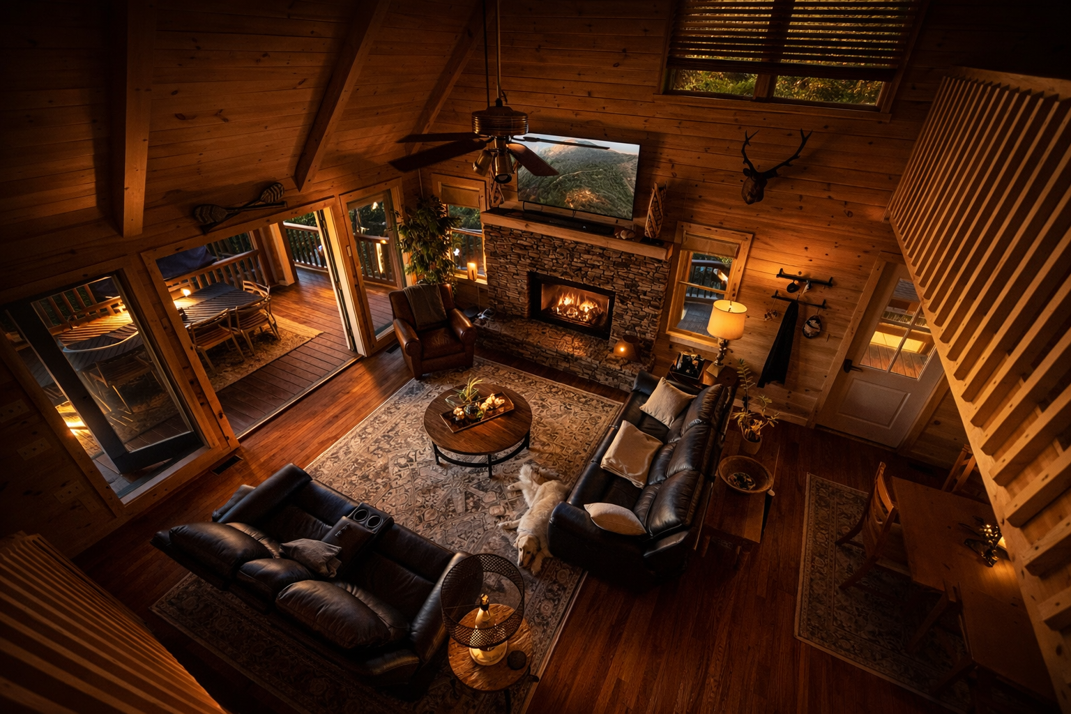 A cozy living room with wood-paneled walls and high ceiling, featuring a stone fireplace, large television, and leather sofas. There are windows and a door leading to an outdoor deck with seating, and warm lighting enhances the inviting atmosphere. A dog is lying on the rug near the sofas.