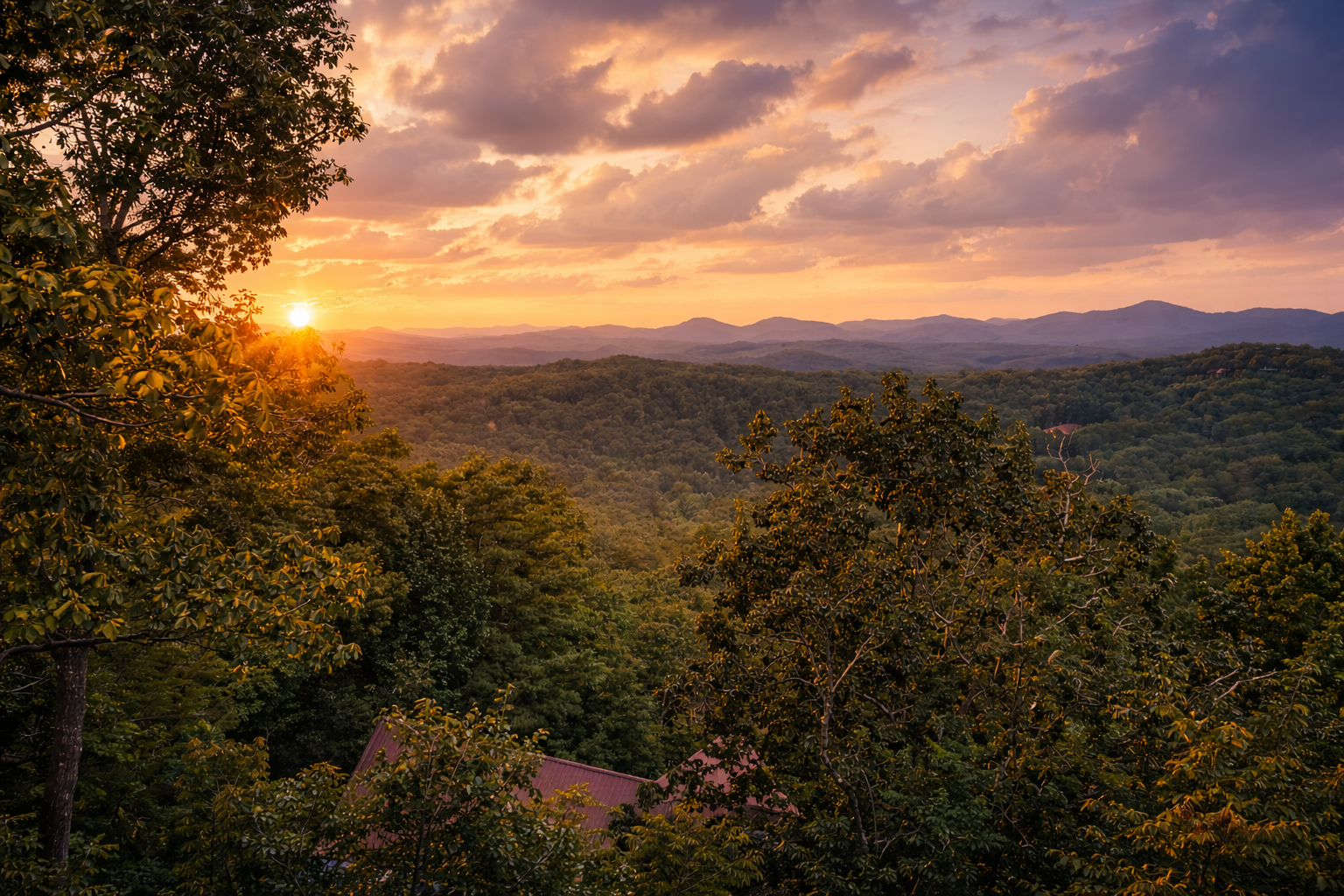 Sunset over a green valley with rolling hills and mountains in the distance, partly cloudy sky, trees in the foreground, and rooftops visible among the trees.