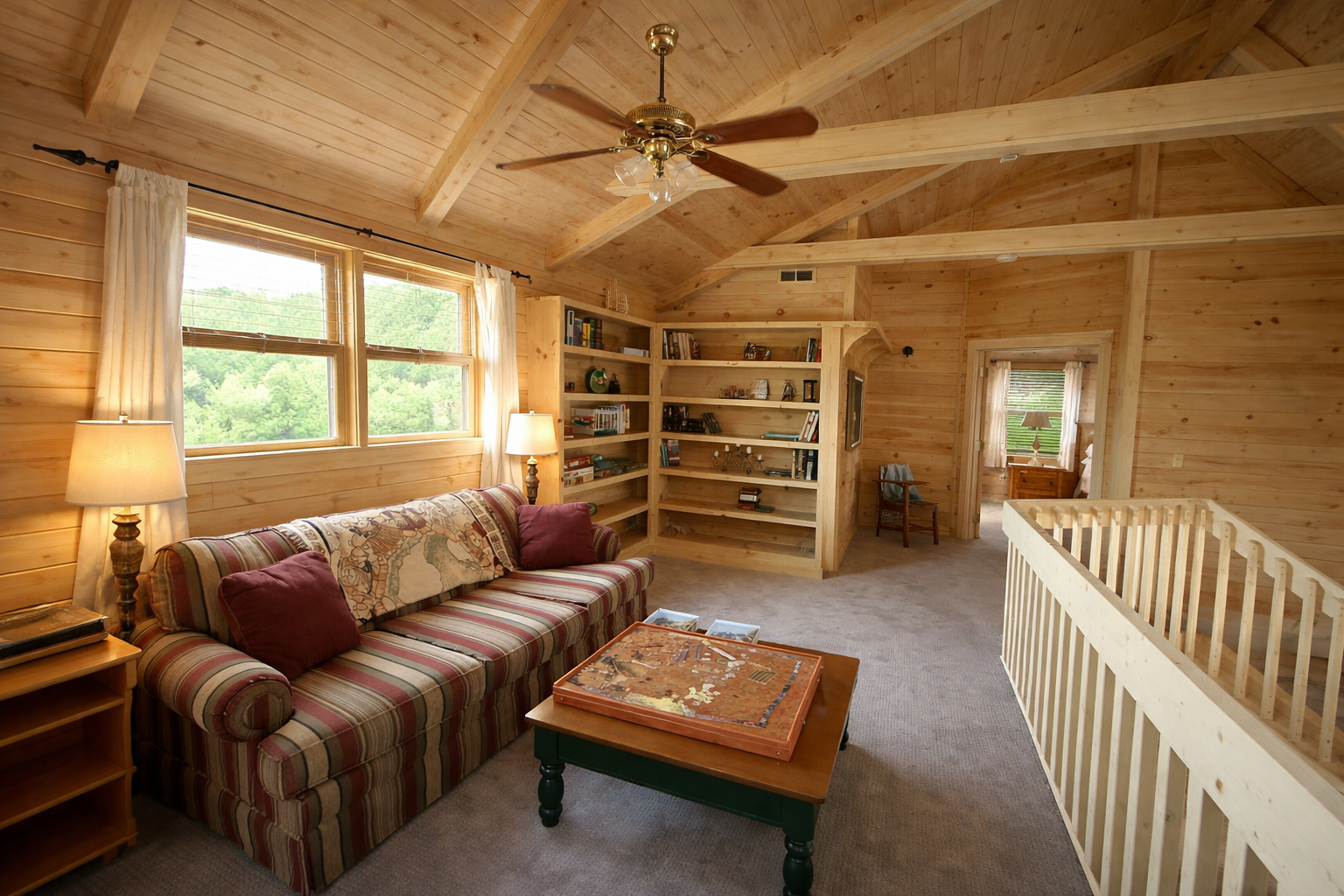 Living room with wooden walls and ceiling, featuring a striped sofa with red pillows, a wooden coffee table with a puzzle, a bookshelf, two lamps, a ceiling fan, and windows with curtains overlooking greenery.