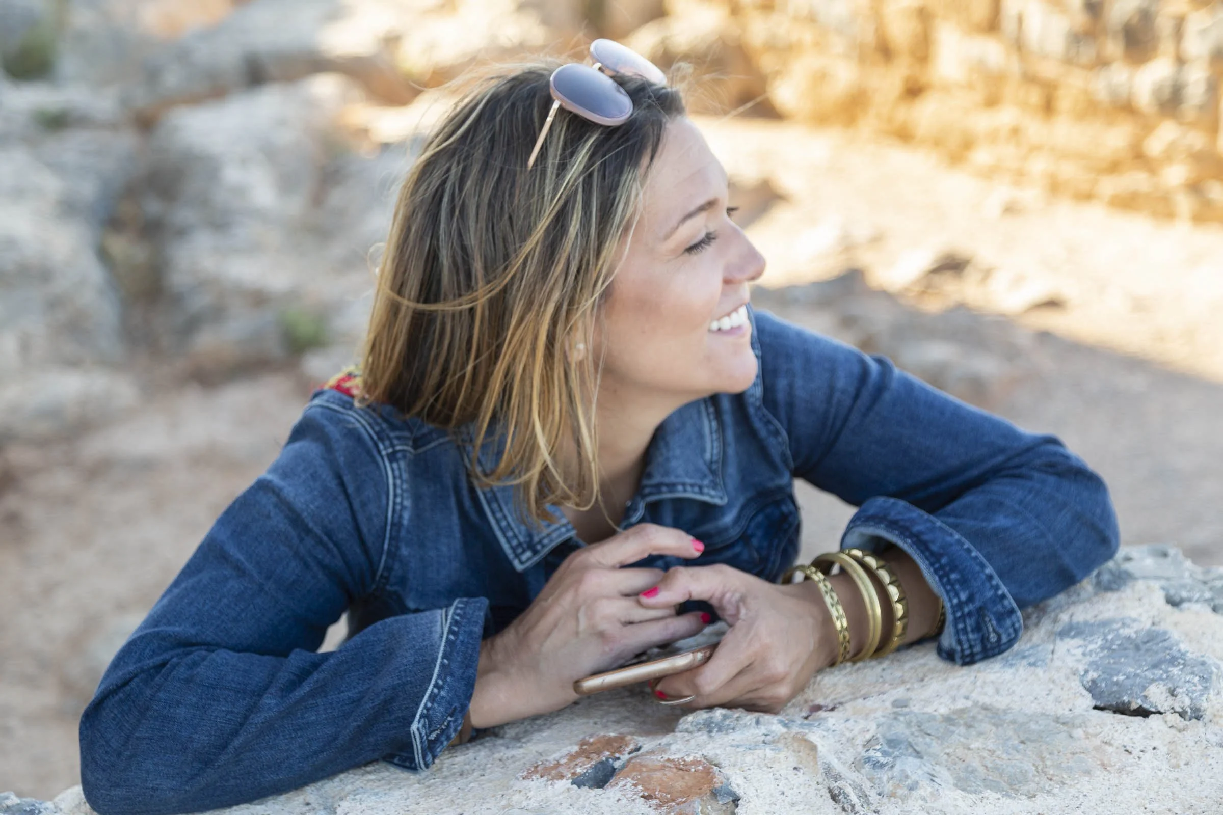 A woman with shoulder-length hair wearing sunglasses on her head, a denim jacket, and gold bracelets is smiling and leaning on a rock outdoors in a natural setting with rocks and trees in the background.