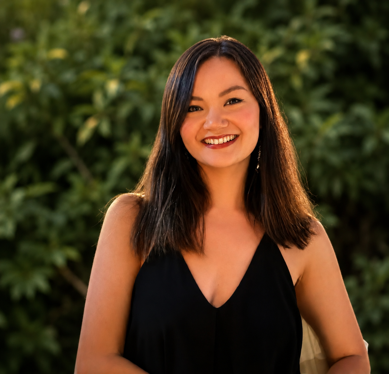 A young woman with shoulder-length dark hair, smiling, wearing a black sleeveless top, standing outdoors with green foliage in the background.