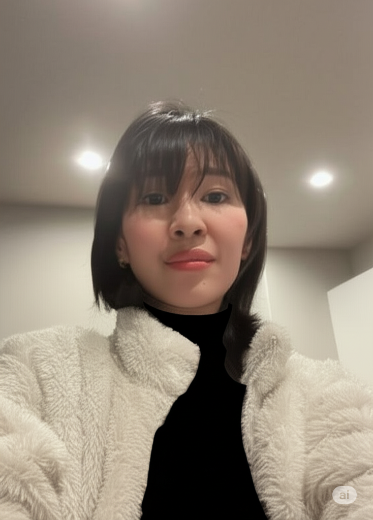 A woman with short black hair, wearing a beige fuzzy jacket over a black top, looking at the camera with a slight smile in a well-lit room.