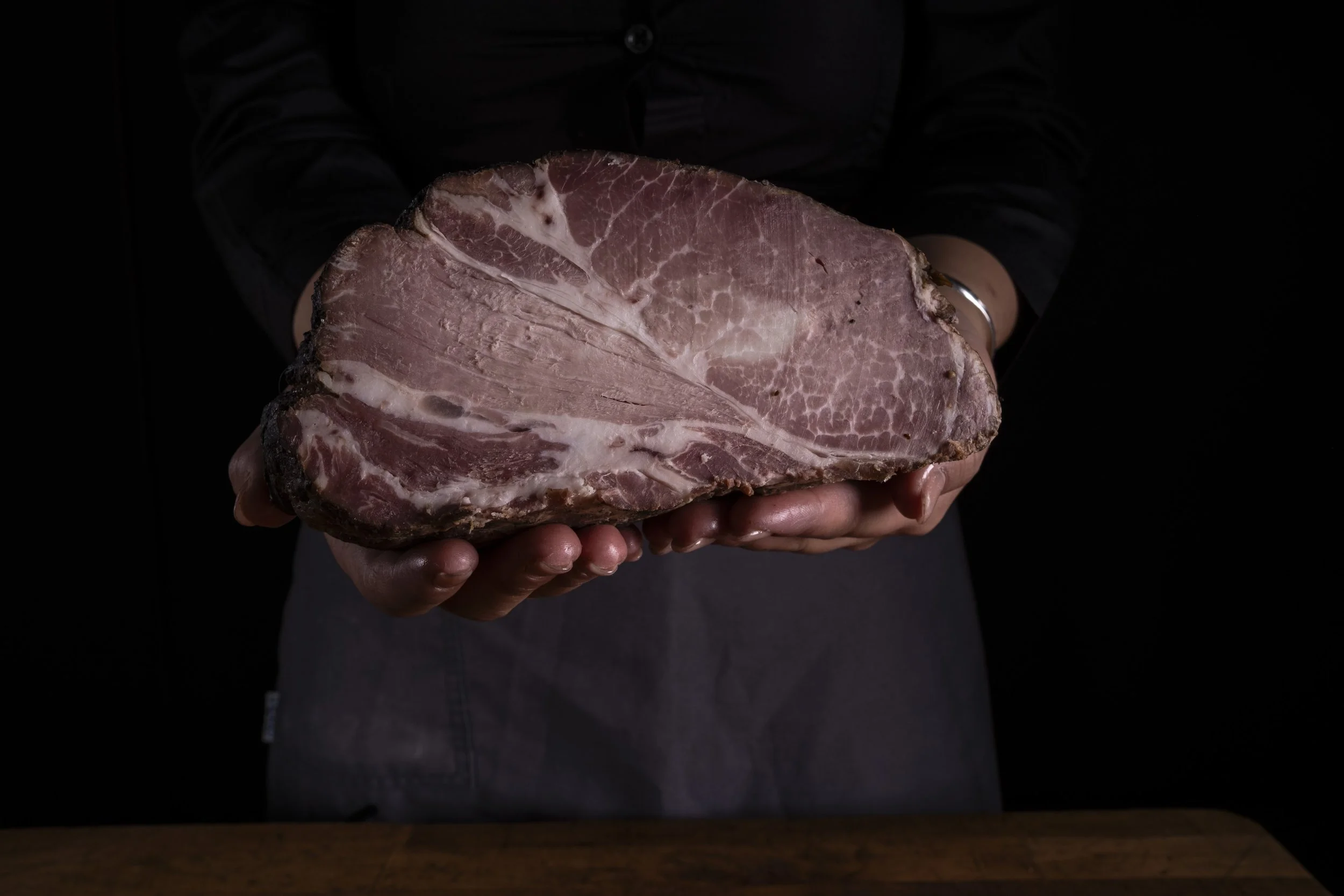 Una persona tiene un grande pez arrosto rosa con bordi più scuri, tenuto con entrambe le mani sopra un tavolo di legno, sullo sfondo uno sfondo nero.