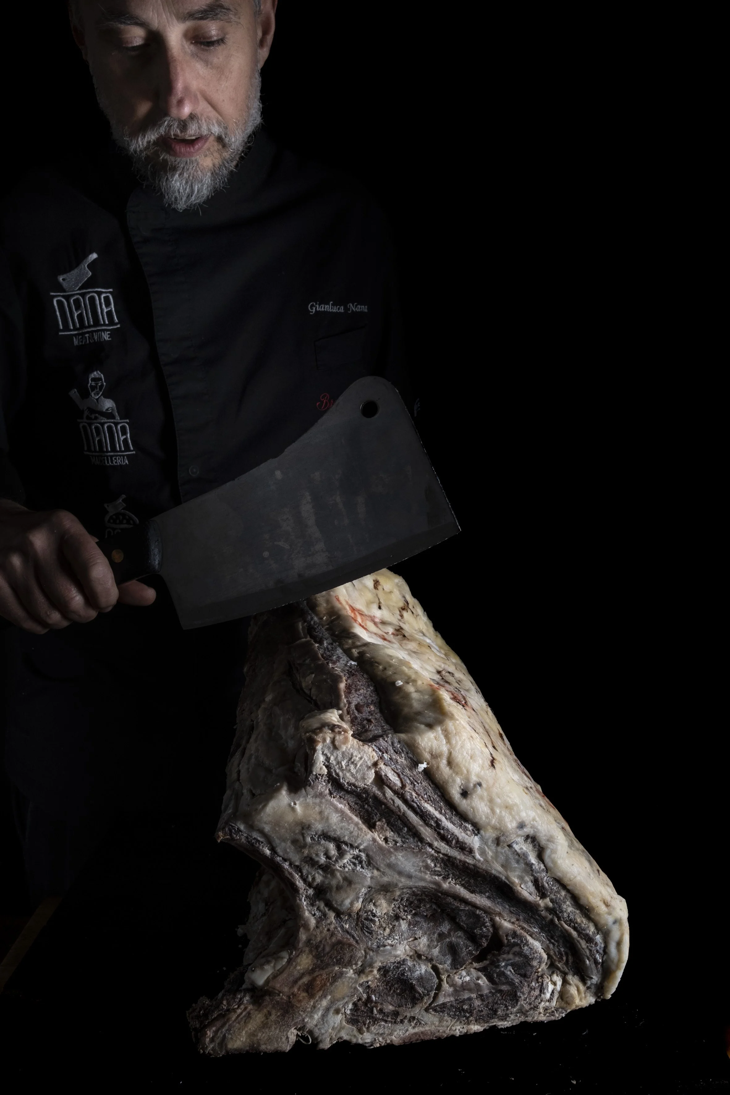 Un uomo con barba che indossa un giacca da chef taglia un grande pezzo di carne con un coltello.