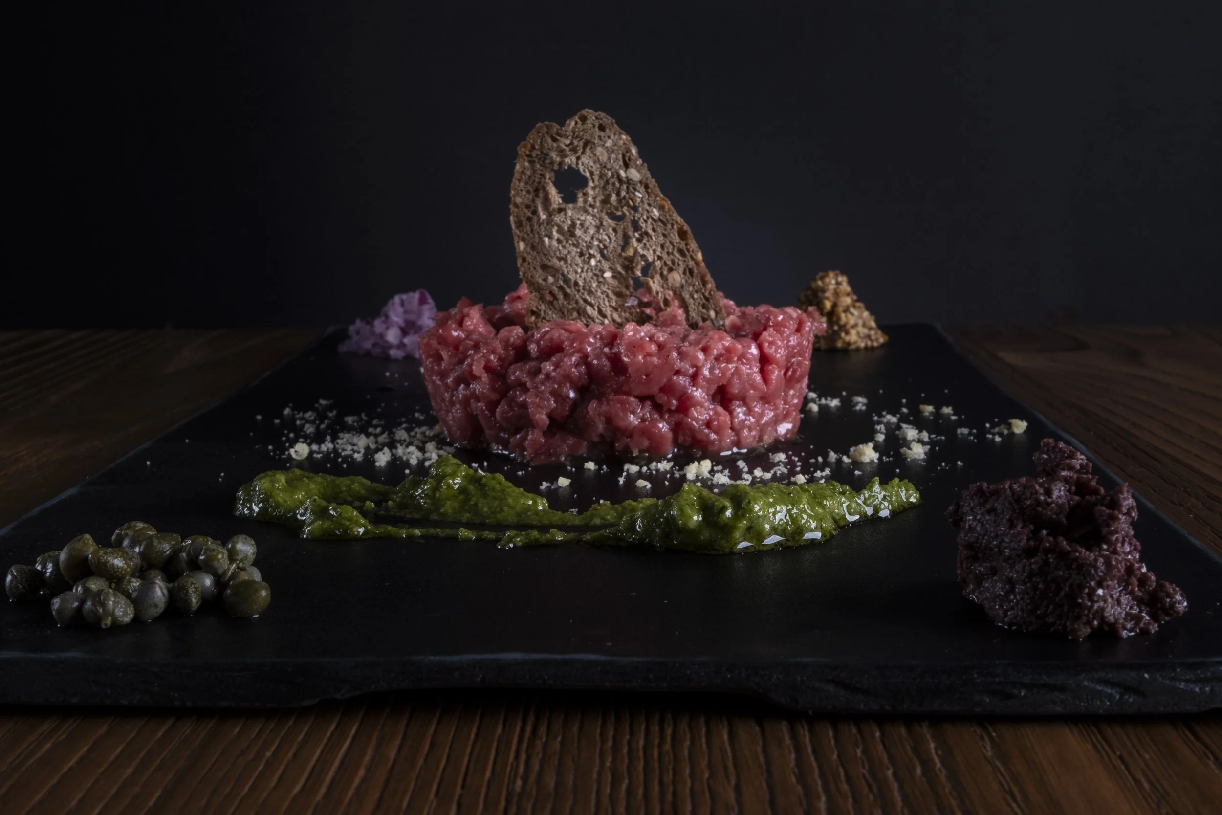 Piatti di carpaccio di carne con vari accompagnamenti, tra cui capperi, salsa verde, burro di olive, e una fetta di pane croccante, serviti su un piatto nero