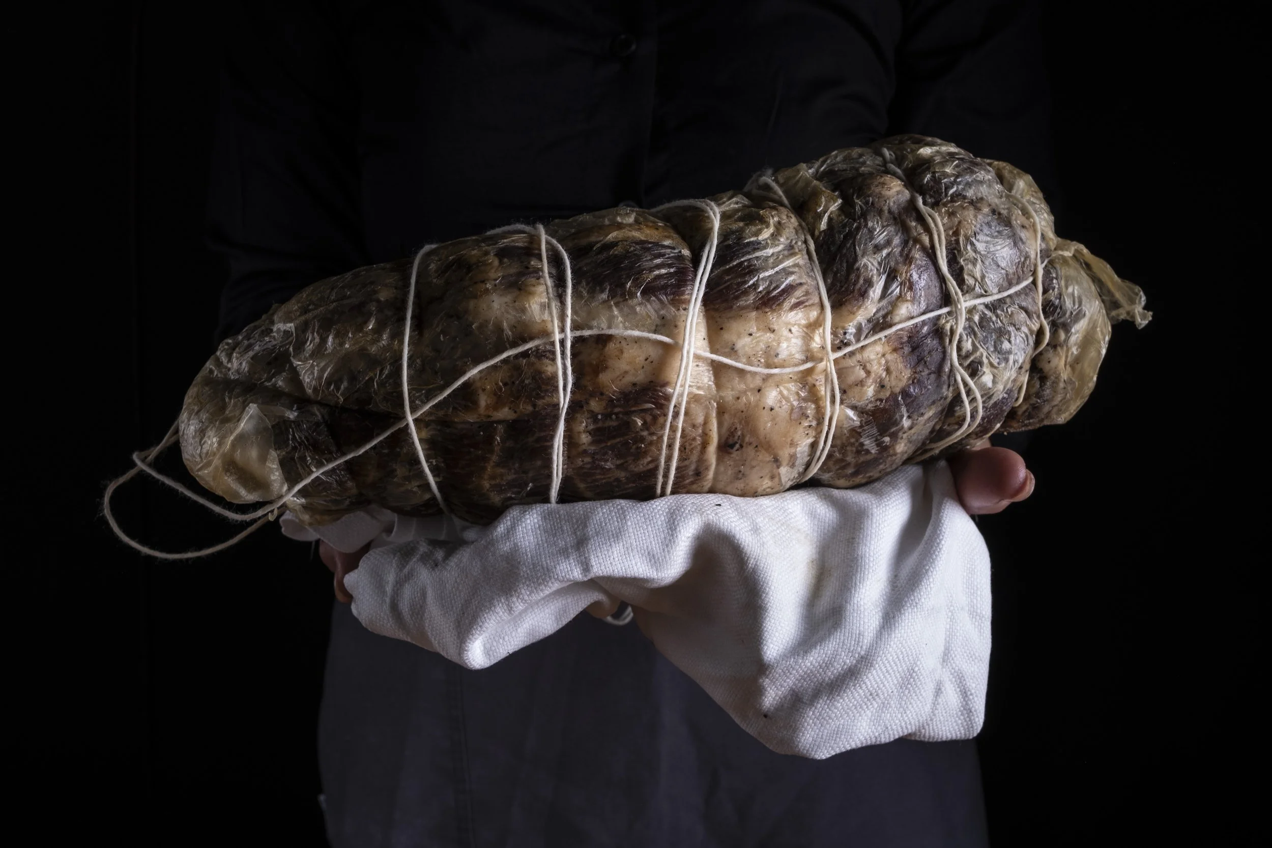 Persona che tiene una grande forma di salume avvolta in plastica e legata con spaghi, posata su un panno bianco.