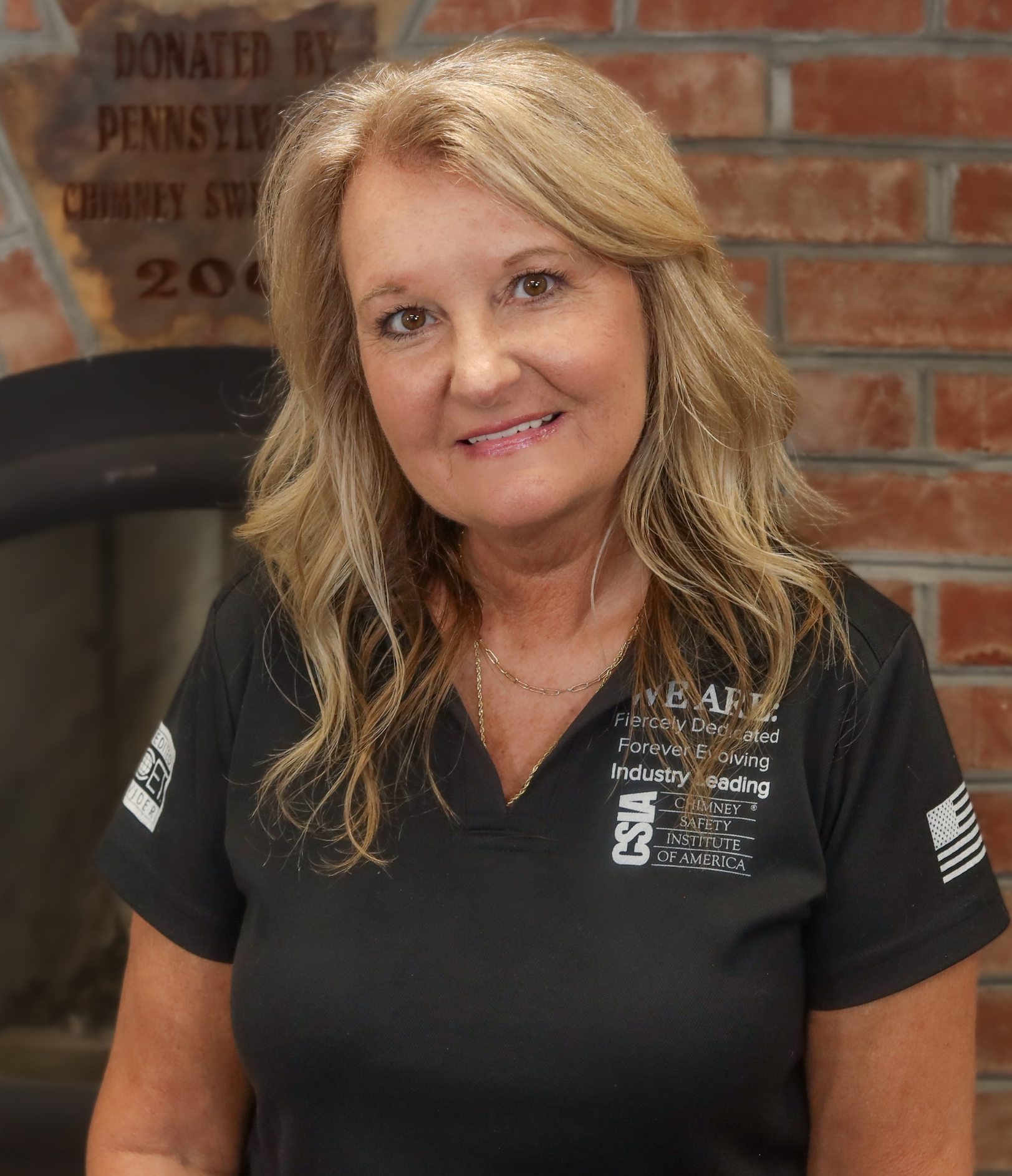 A woman with curly blonde hair smiling, wearing a pink shirt with a logo and gold necklace, standing in front of a brick wall.