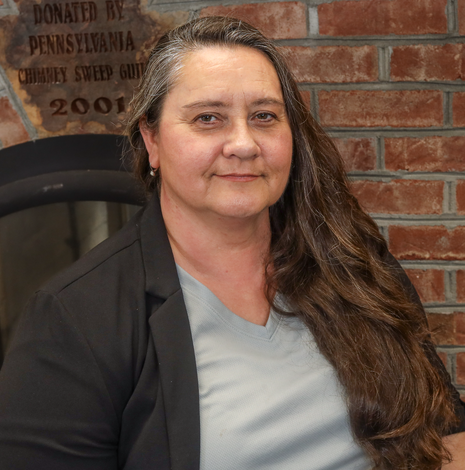 A woman with glasses and long brown hair in a braid, wearing a black collared shirt with white text, standing in front of a brick and stone wall.