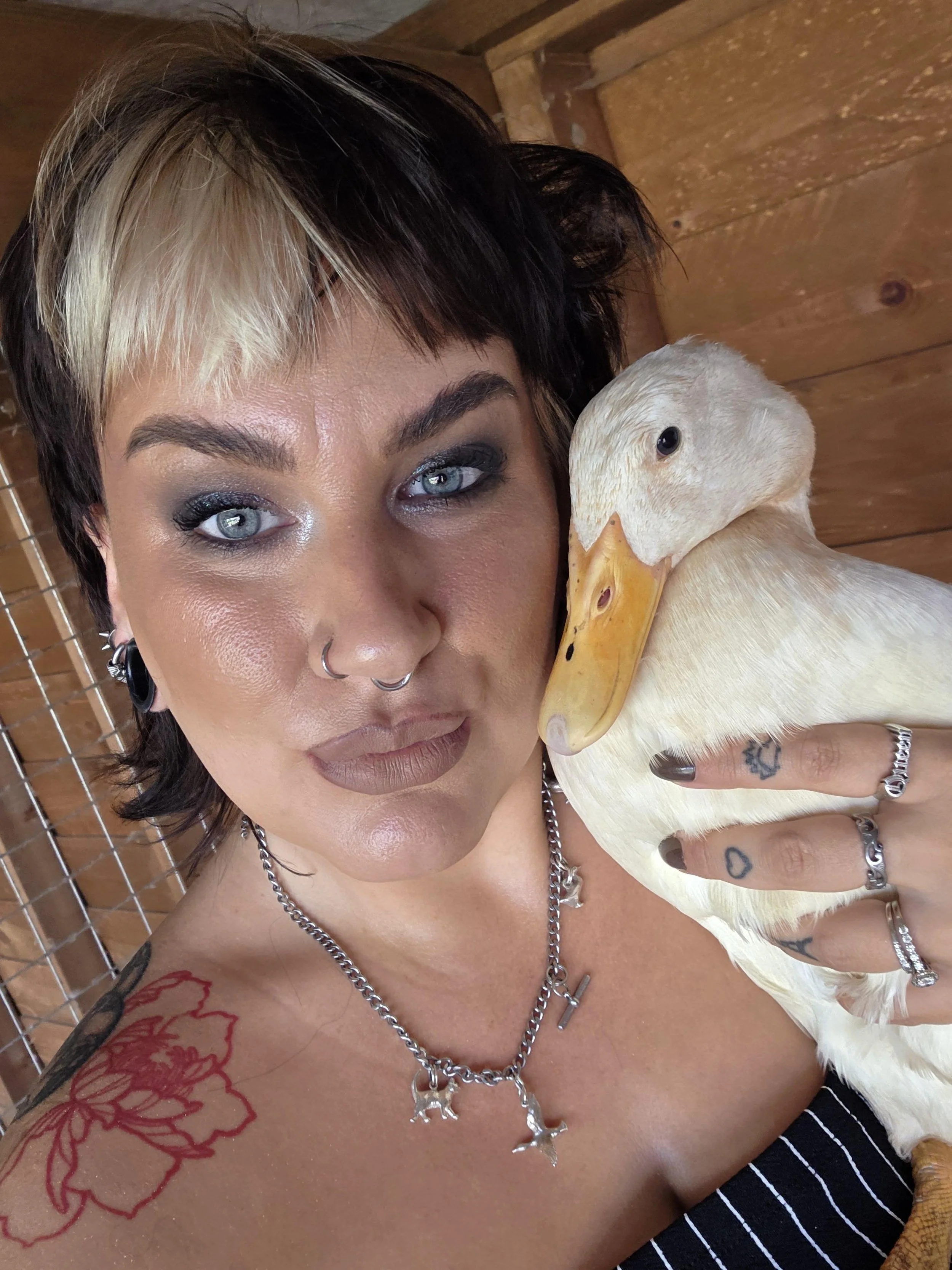 A woman with short, two-toned hair holding a white goose close to her face, with tattoos visible on her shoulder and hand, and wearing jewelry, in a wooden setting.