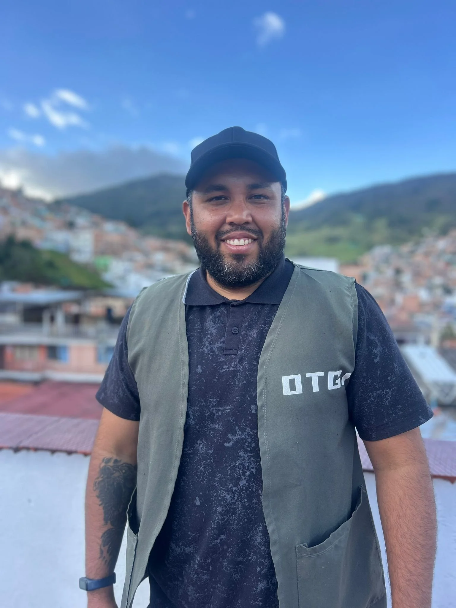A man standing outdoors with a hilly town and blue sky in the background, smiling at the camera.