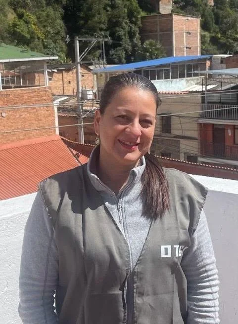 Woman standing outdoors on a rooftop with buildings and trees in the background, wearing a gray vest over a light gray long-sleeve shirt, smiling at the camera.