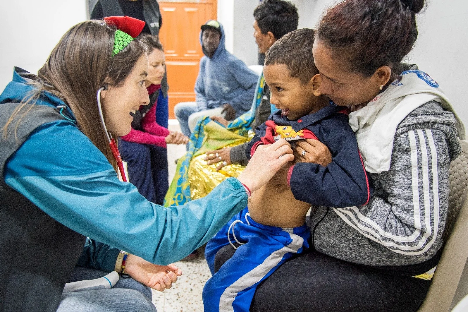 A young woman in a blue jacket using a stethoscope to check a young boy's torso while another woman and boy sit nearby in a waiting area.