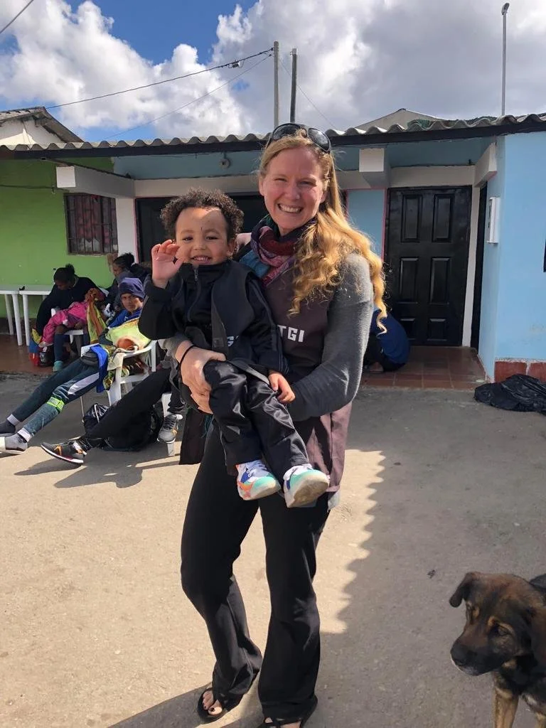 A woman holding a young child, both smiling, outdoors in front of colorful buildings with other children sitting in the background and a dog nearby.