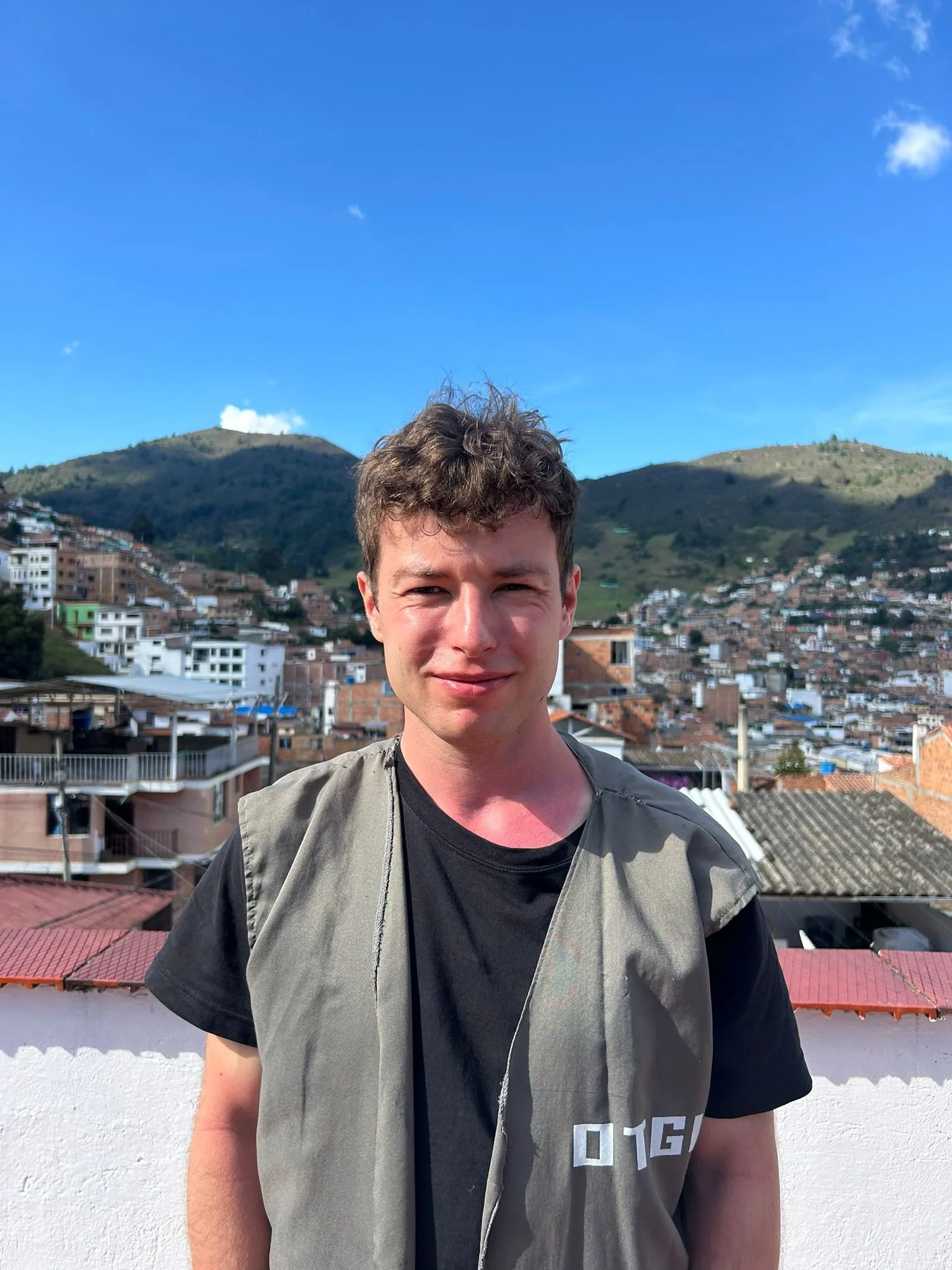 A young man with short curly brown hair standing outdoors with a sunny blue sky and a hillside filled with houses in the background.