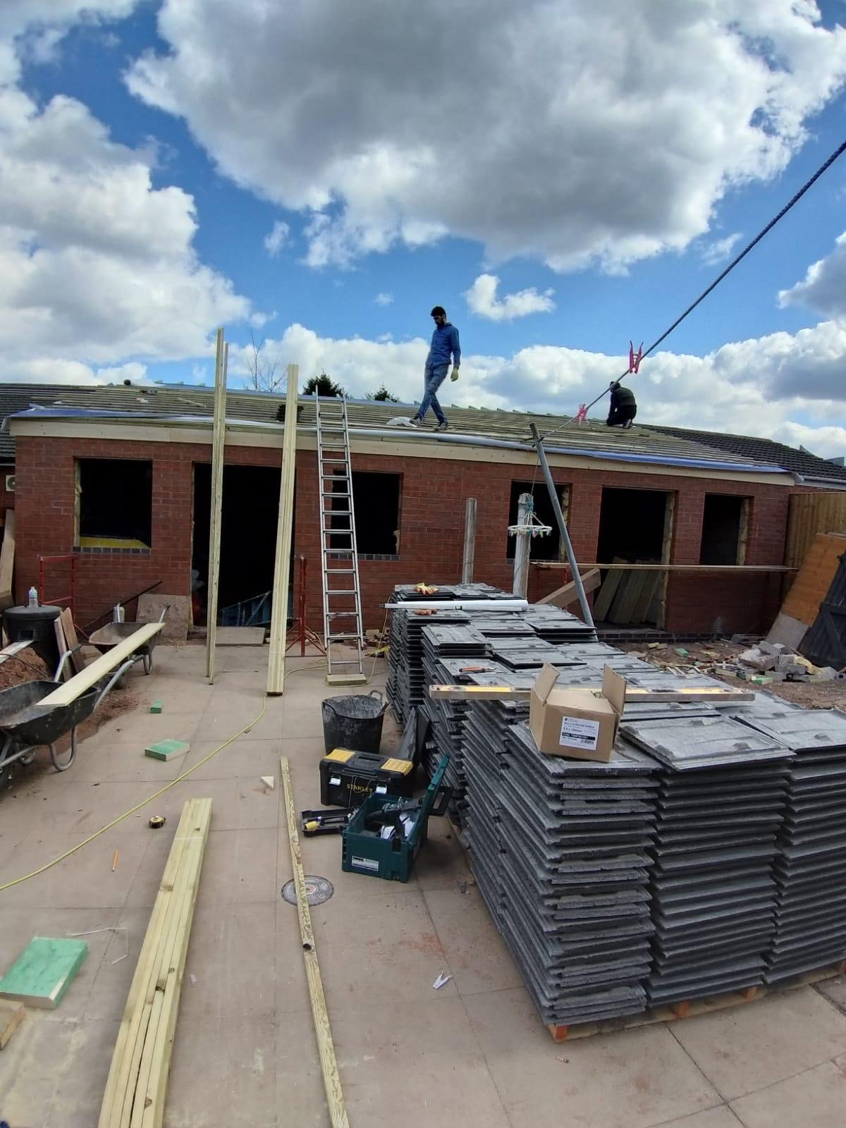 Construction workers installing roofing on a house under a cloudy sky. Building materials and tools are scattered around a concrete patio.