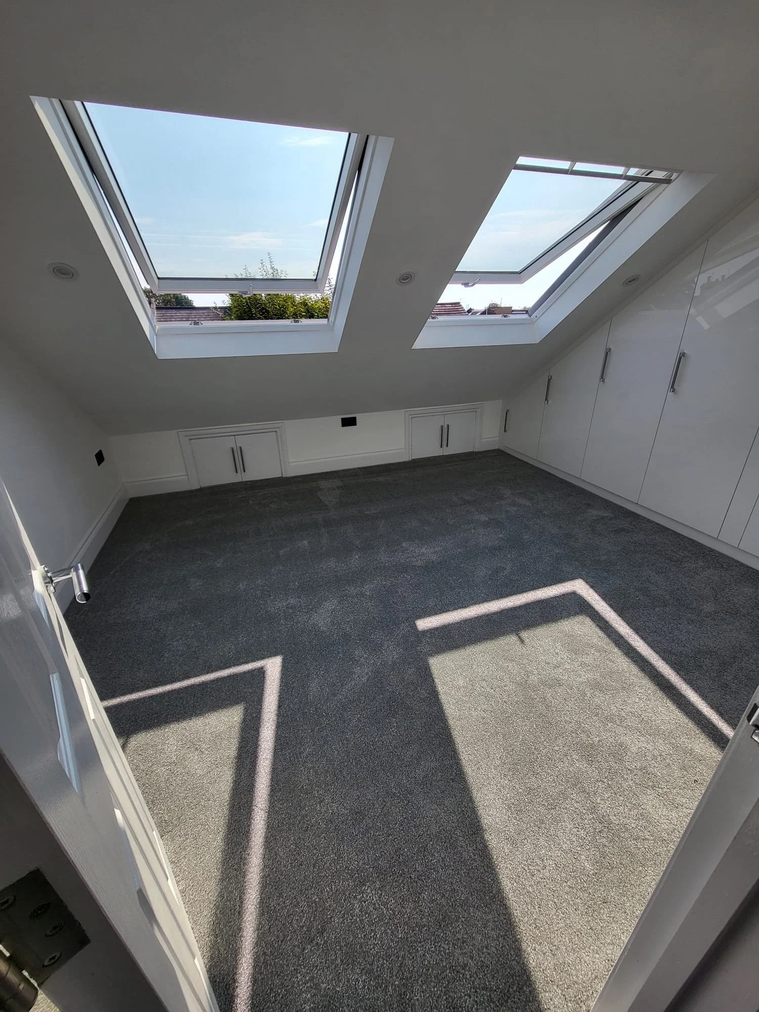Empty attic room with two large skylight windows, white built-in cabinets, and a gray carpeted floor.