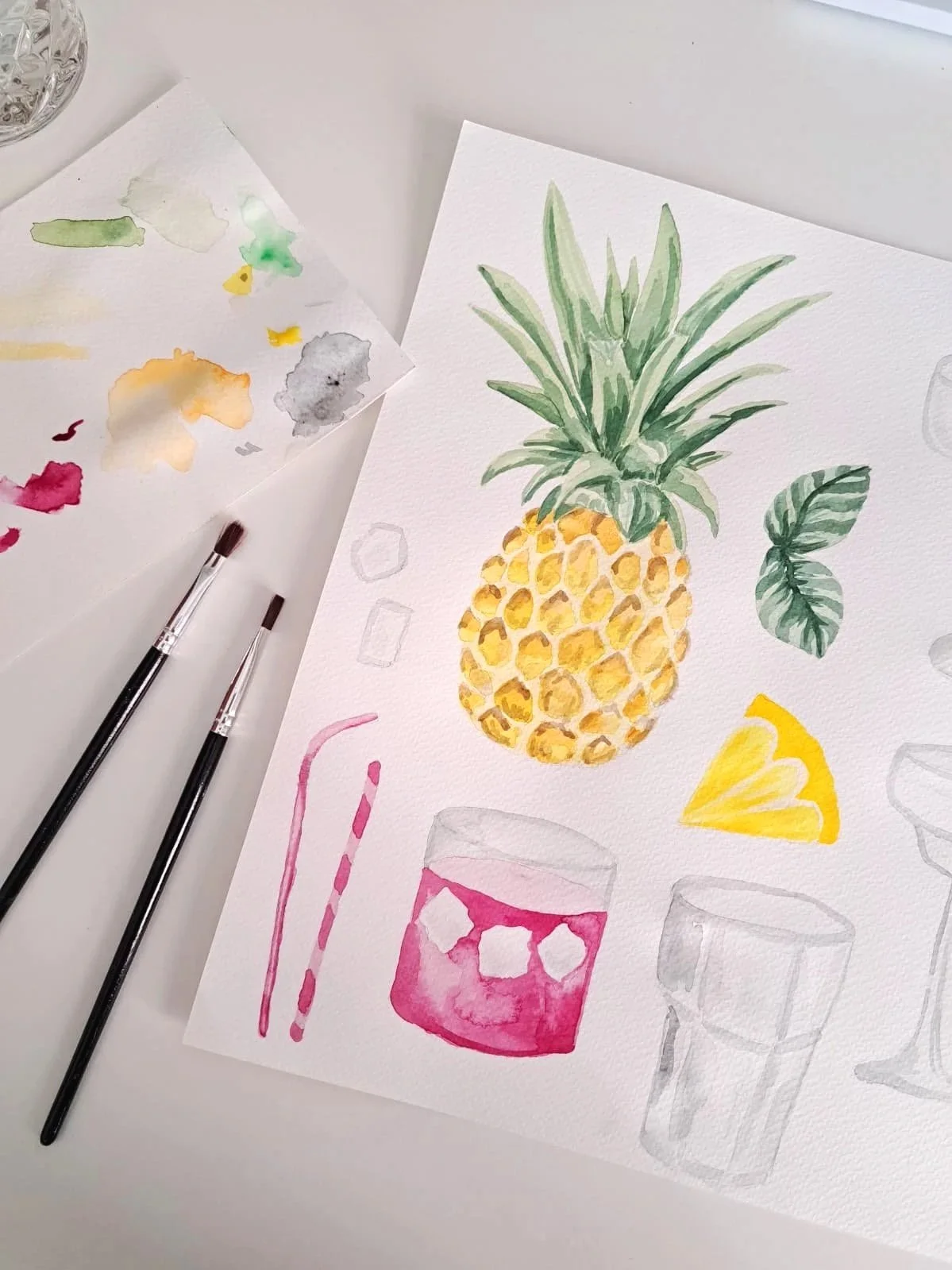 A watercolor painting on paper featuring a pineapple, a slice of lemon, a pink cocktail glass with ice, and an empty clear glass, with watercolor paints and brushes nearby.