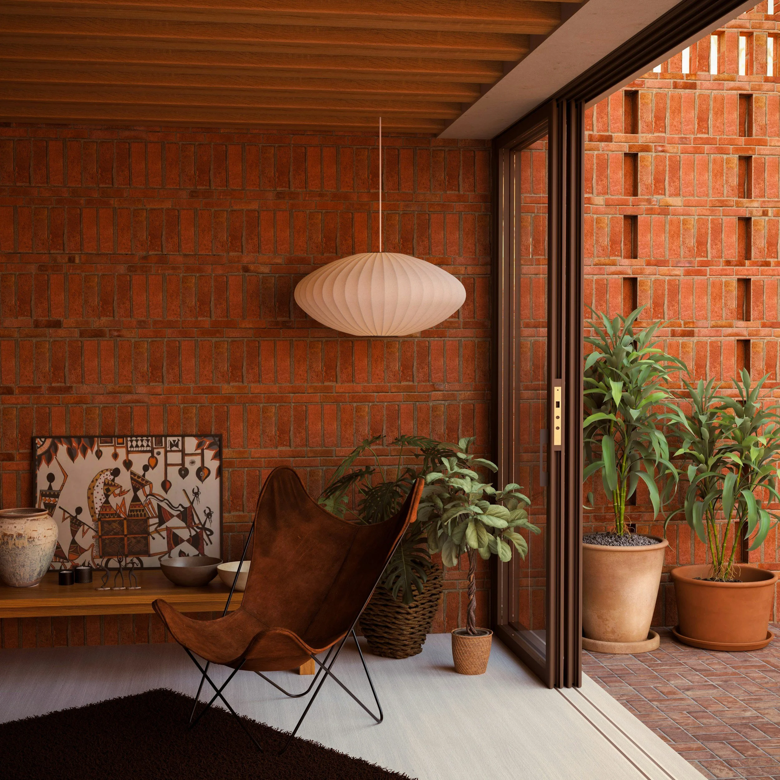 Interior view of a room with a brick wall, wooden ceiling, and glass sliding door leading to a balcony with large potted plants. There is a hanging white pendant light, a brown butterfly chair, a low wooden table with decorative items, and a patterned painting leaning against the brick wall.