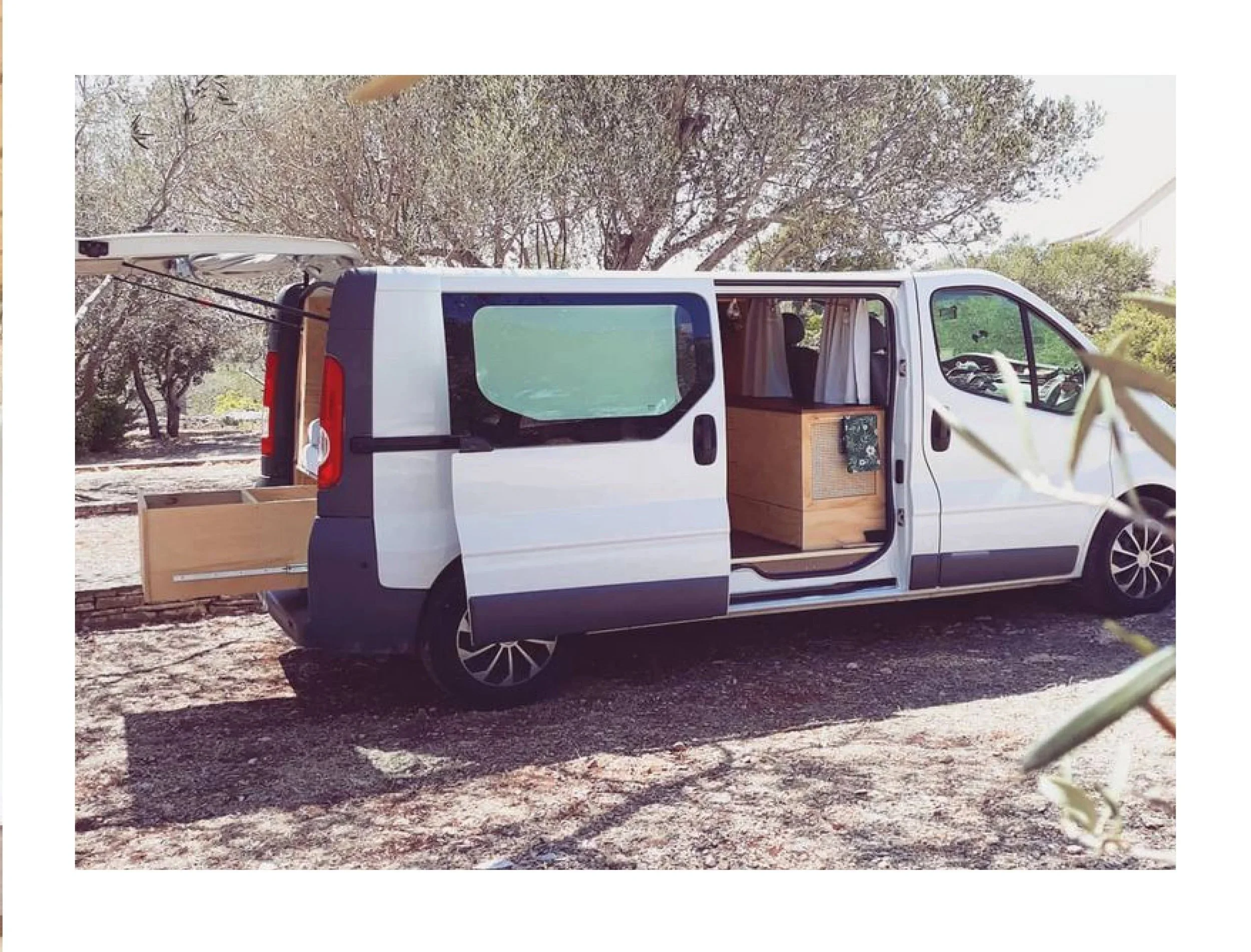 A white van with open sliding door showing wooden interior furniture, parked outdoors among trees and bushes, with the back door and side open.