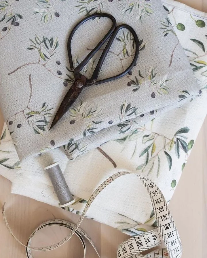 A pair of sharp scissors, a spool of gray thread, and a white measuring tape on a floral fabric with an olive branch pattern.