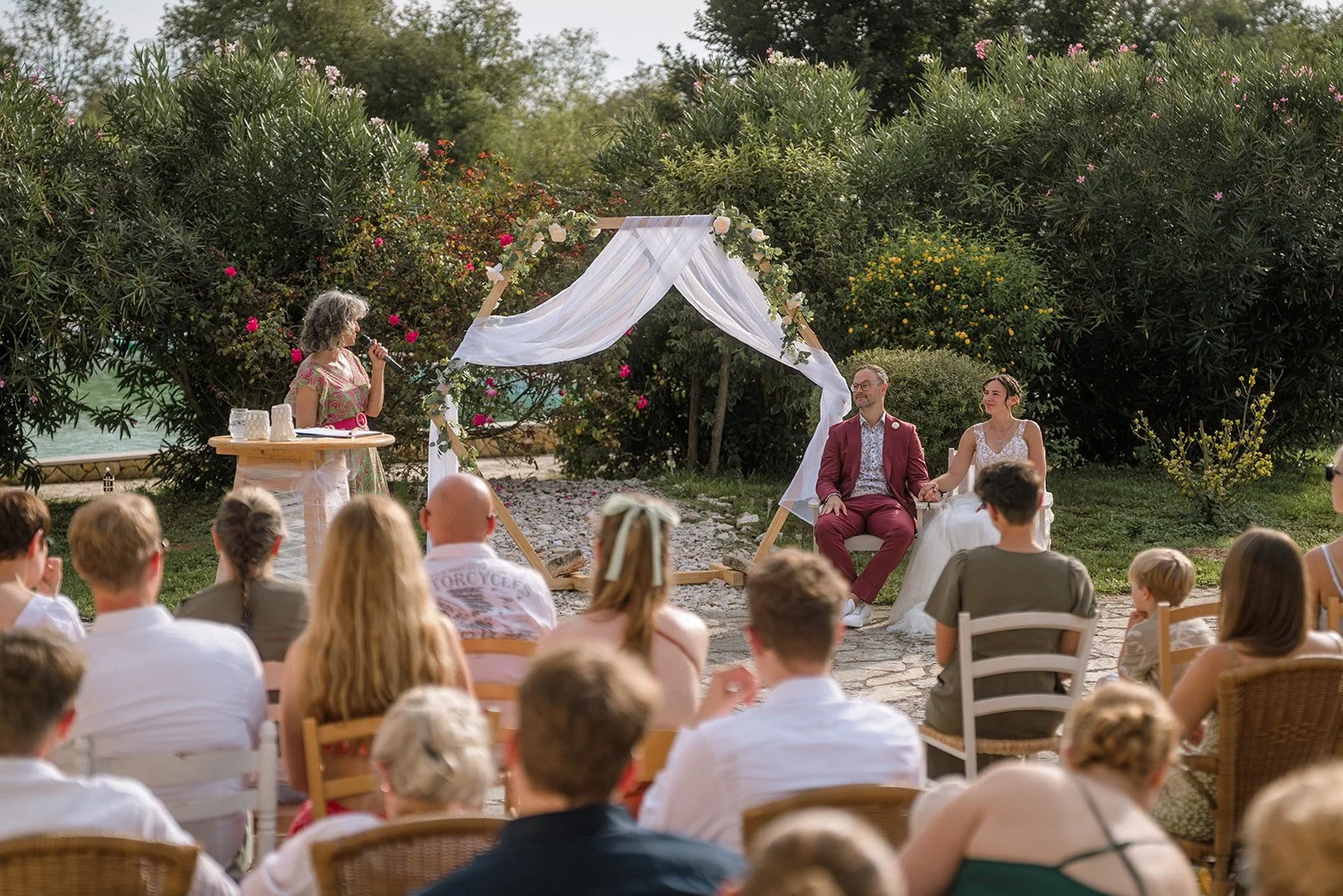 A wedding ceremony outdoors with a bride and groom sitting under a white draped arch decorated with flowers, officiant speaking, seated guests watching, and lush greenery in the background.