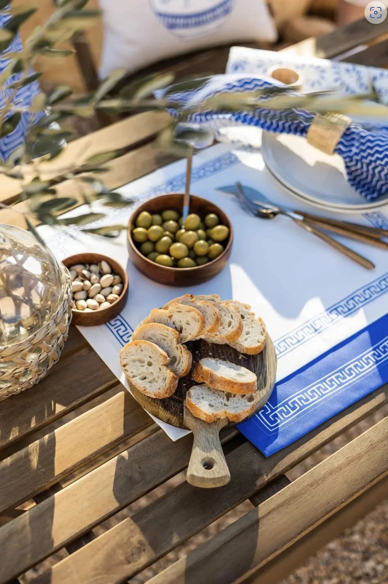 A wooden table set with sliced bread on a wooden paddle, bowls of green olives and pistachios, silverware, a white plate with a blue patterned napkin, and decorative olive branches.