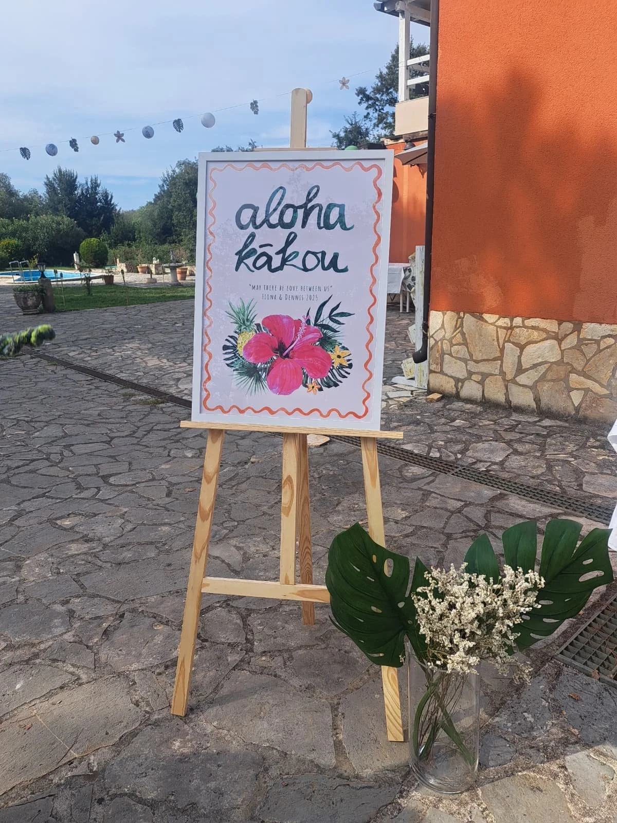 An outdoor sign on an easel with colorful tropical flowers and the words 'Aloha Kakou' and a quote, with a vase of white flowers and large green leaves in front. In the background, trees, a pool, and a building are visible.
