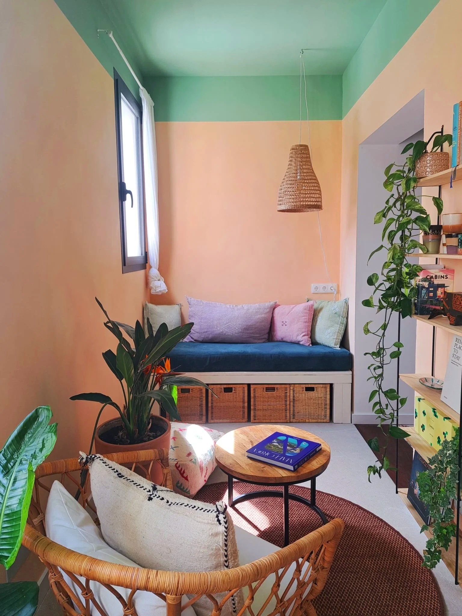 Cozy living room with pastel-colored walls, a window with white curtains, a built-in bench with cushions, a small round wooden coffee table with a book, a rattan armchair with pillows, a large potted plant, and an open shelf with books and decor.
