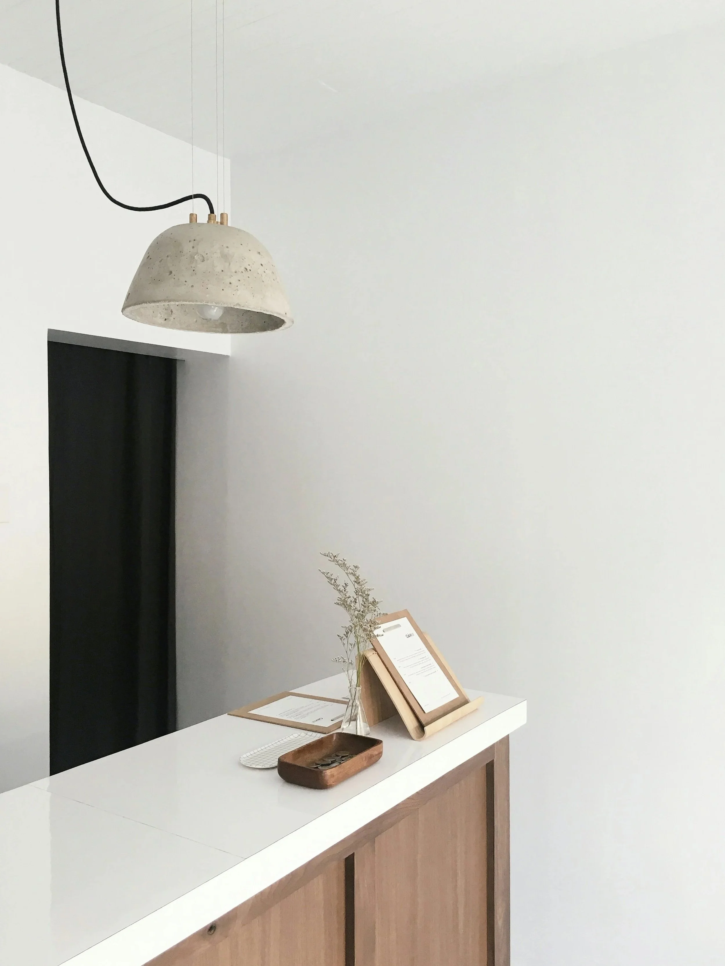 Minimalist reception area with a white countertop, a wooden panel, a small vase with dried plant, a metal tray, and framed documents. Hanging concrete pendant lamp above.