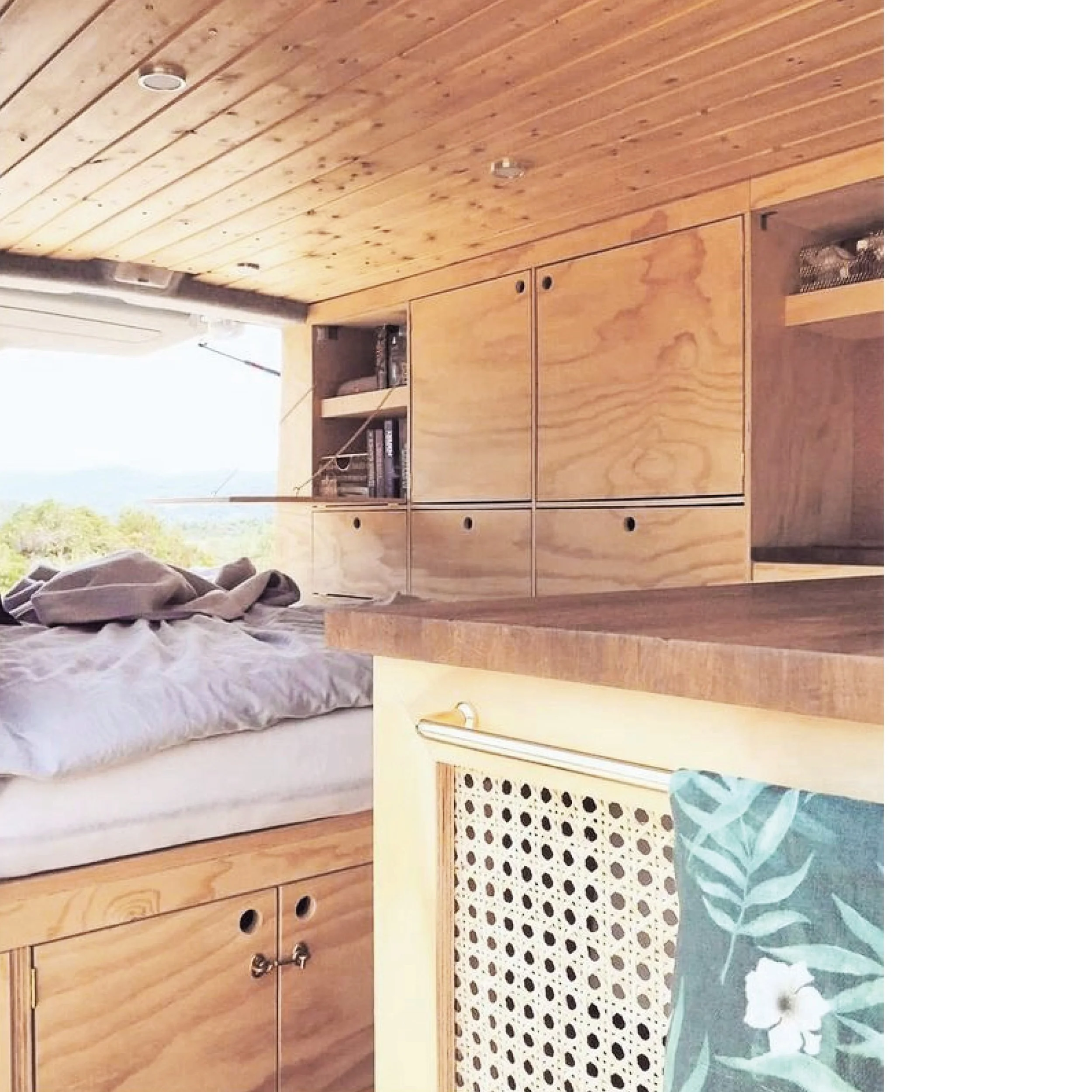 Interior of a wooden camper or tiny home with cabinets, a bed with wrinkled bedding, and a window showing an outdoor landscape.