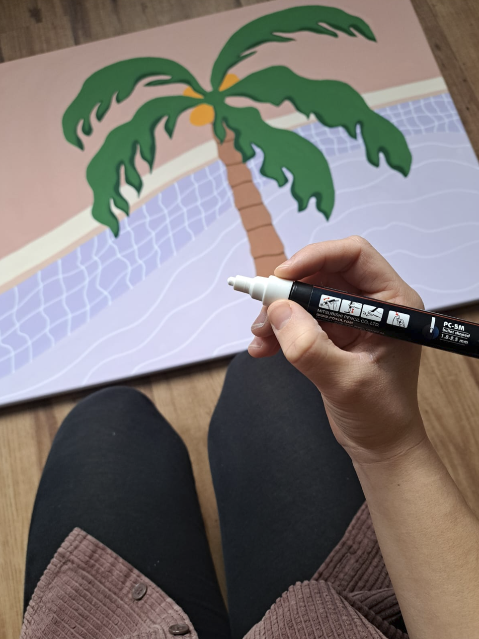 Person drawing a tropical scene poster of a palm tree with green leaves and brown trunk, with a pink sky and a swimming pool on the ground, using a black marker.