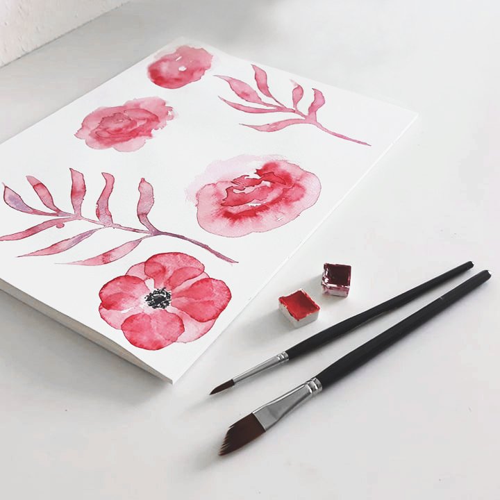 Watercolor painting of pink flowers and leaves on white paper with two paintbrushes and watercolor paints.