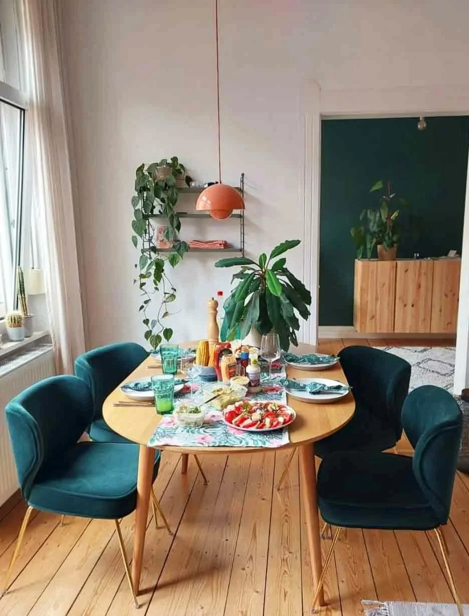 A dining room with a wooden table set for a meal, surrounded by four teal velvet chairs. The table has plates, glasses, condiments, and a pizza. There are plants on the windowsill, a hanging orange lamp, and a large leafy plant in the center. In the background, there is a doorway with a dark green wall and a wooden sideboard with a potted plant.