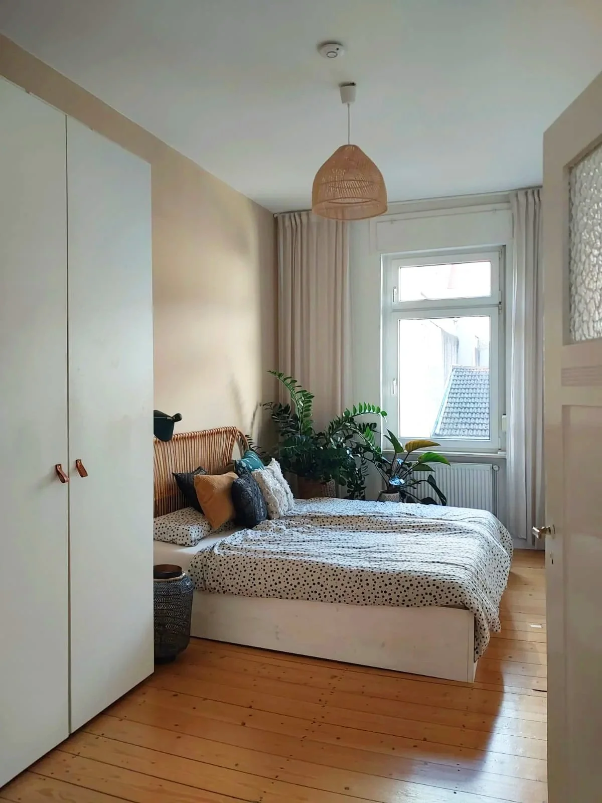 A bedroom with a window, beige walls, a wooden floor, and a white wardrobe. There are plants beside the bed with a headboard, multiple pillows, and a bedspread with a polka dot pattern. A woven lampshade hangs from the ceiling.