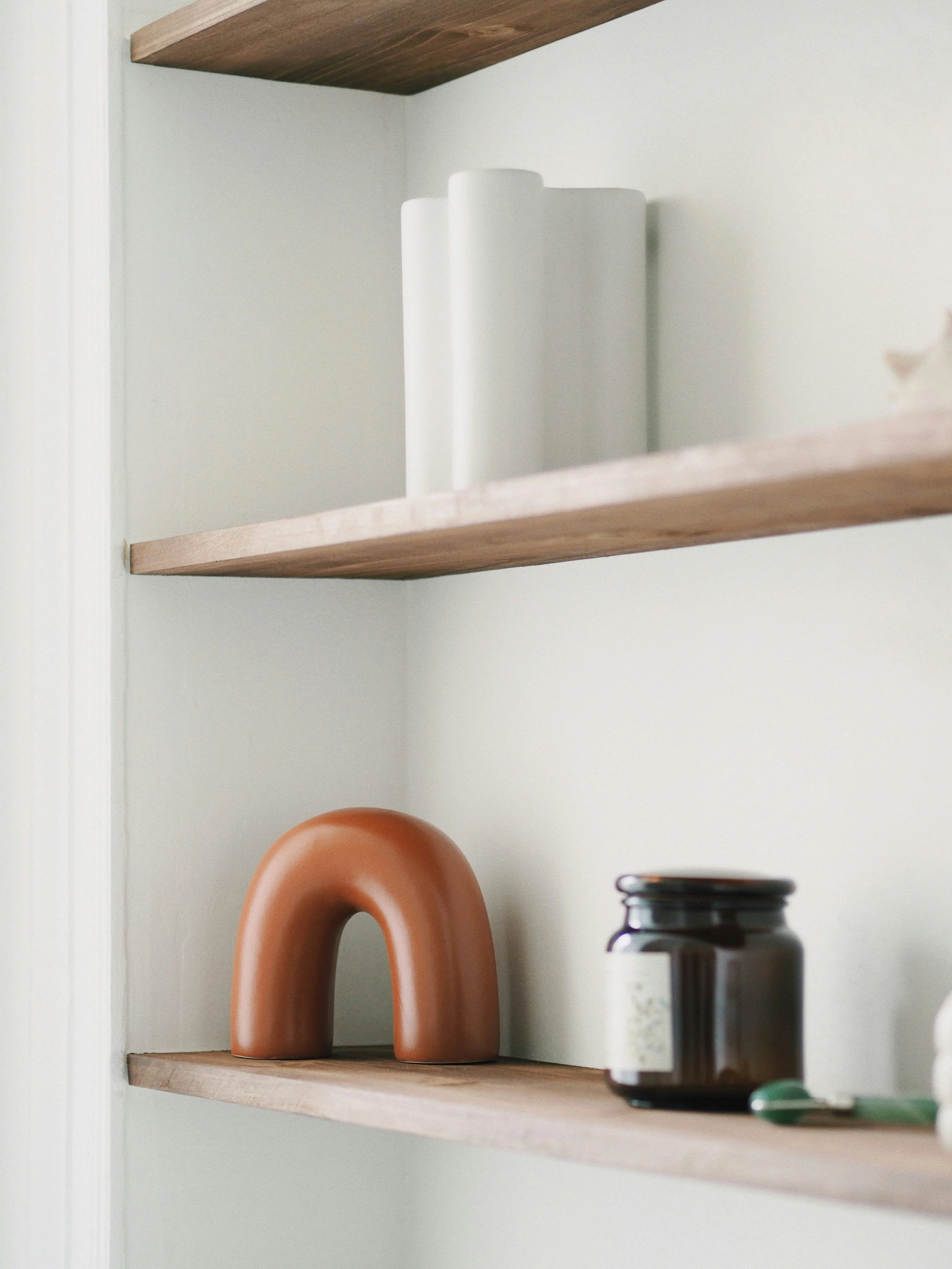 Close-up of wooden shelves with white and terracotta decorative objects, a jar, and a small green item in a room with white walls.