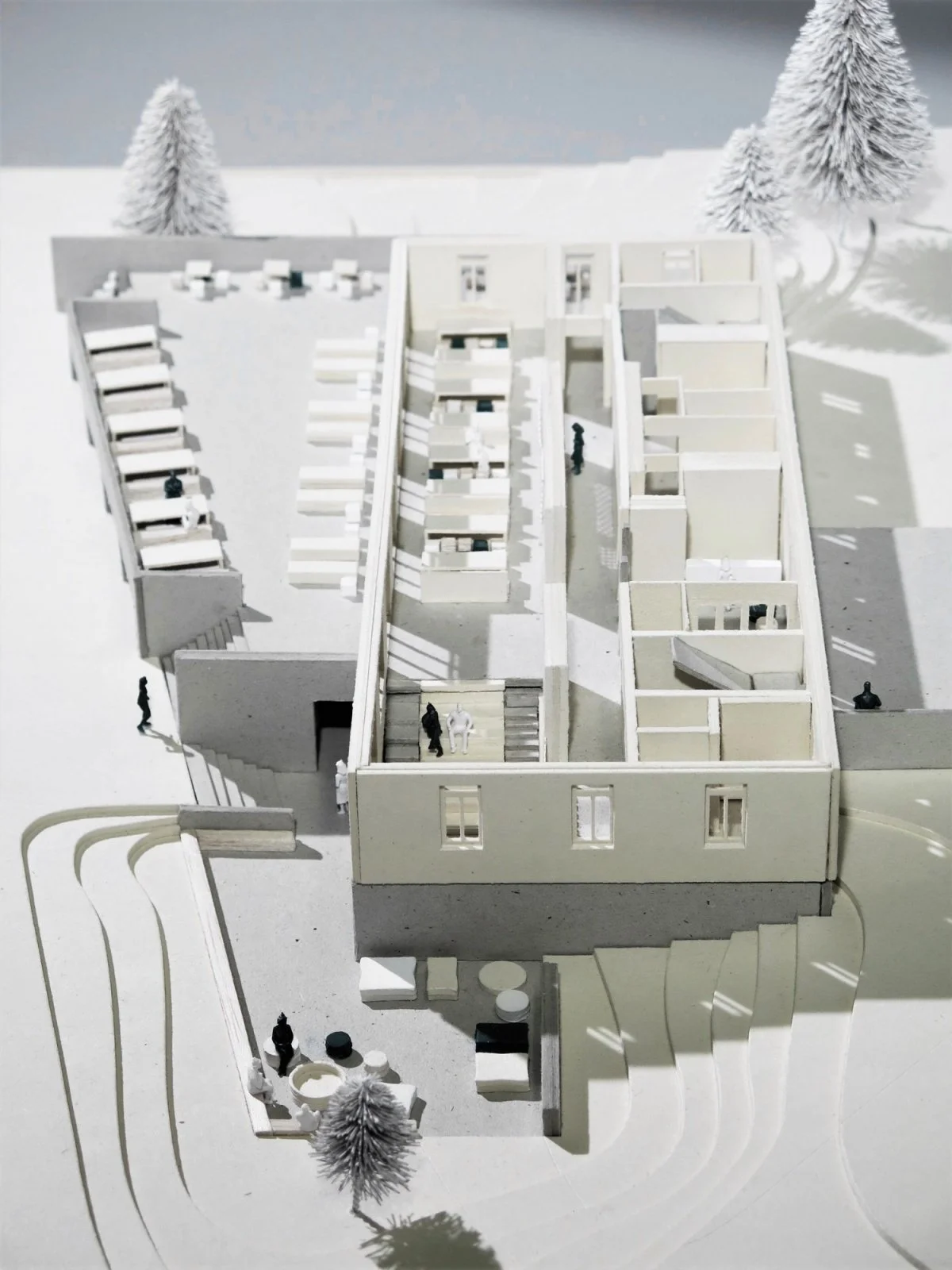 Architectural model of a modern building with multiple floors, exterior and interior walls, and miniature figures representing people, set on a white surface with small model trees in the background.