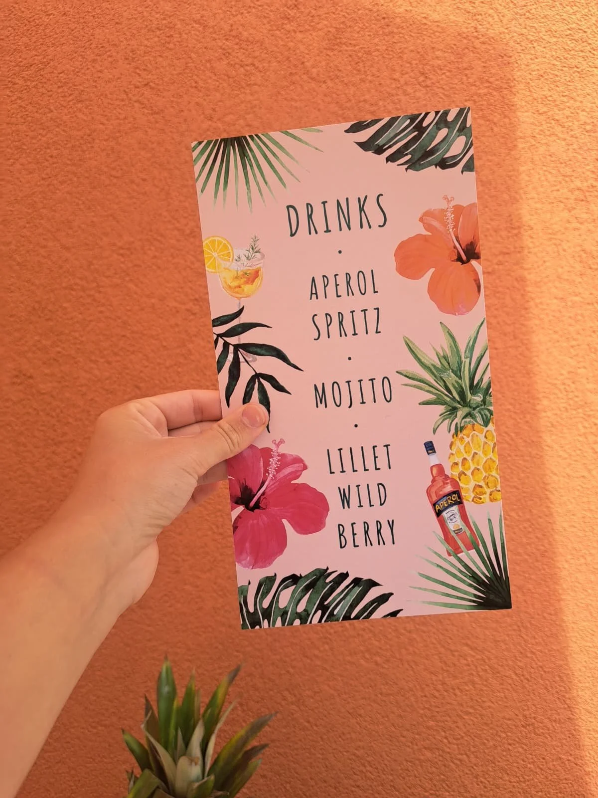 A person holding a pink tropical-themed drink menu with floral and leaf illustrations, listing drinks like Aperol, Spritz, Mojito, Lillet, Wild Berry, against a coral-colored wall.