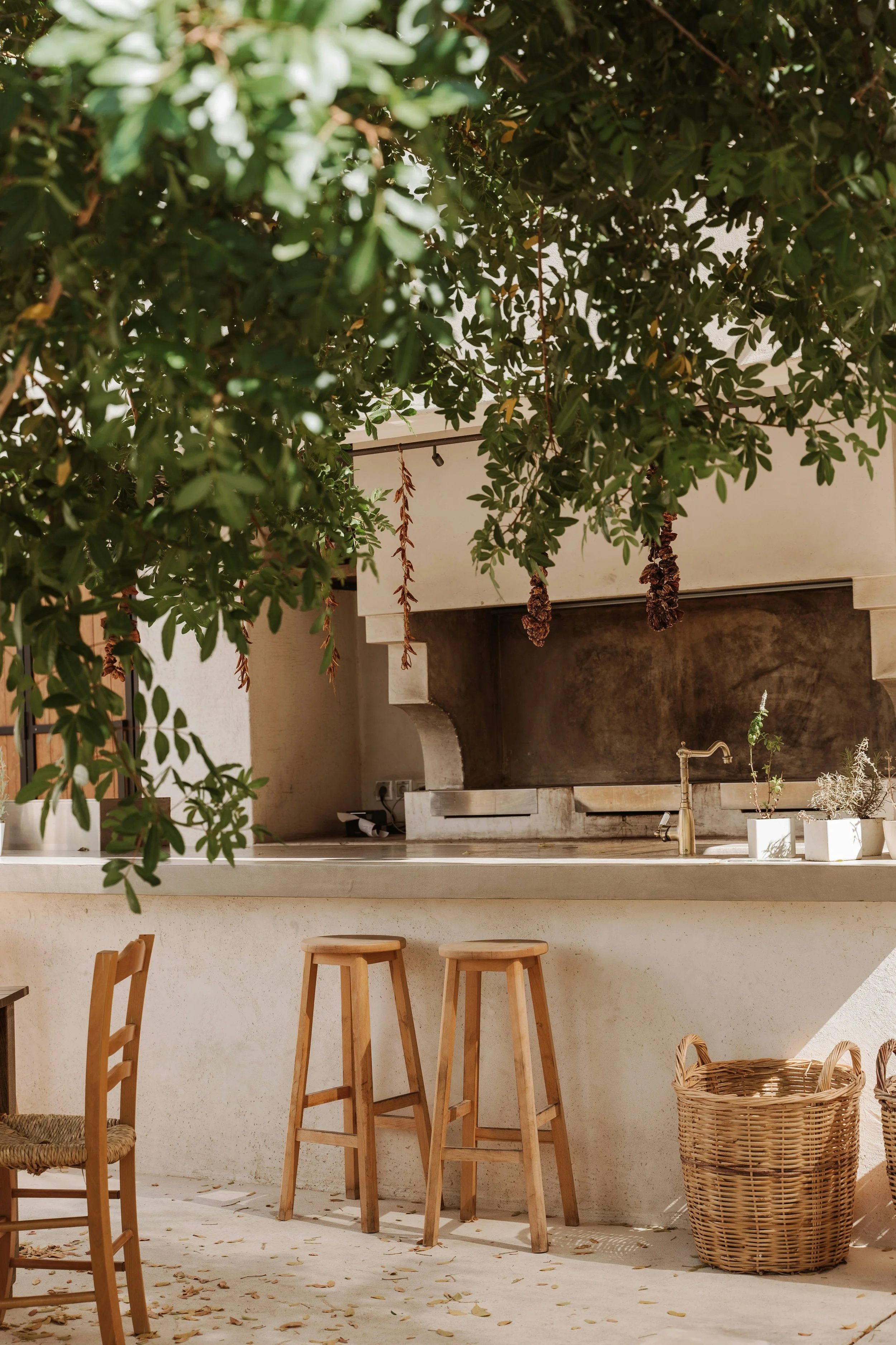 An outdoor kitchen with a white countertop, wooden stools, wicker baskets, and potted plants, partially obscured by overhead leafy tree branches.