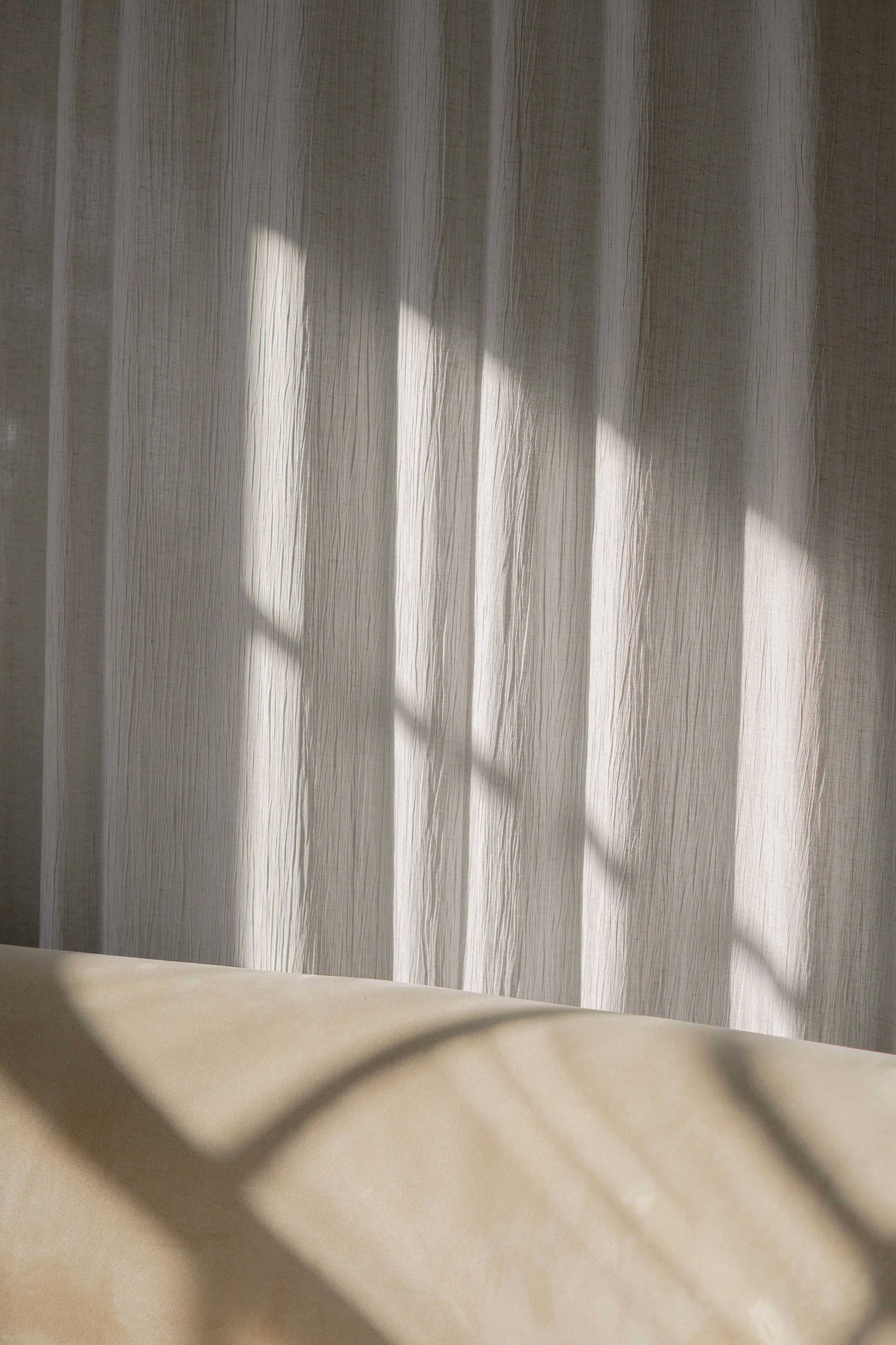 Sunlight streaming through a thin, textured curtain casting shadows on a beige surface.