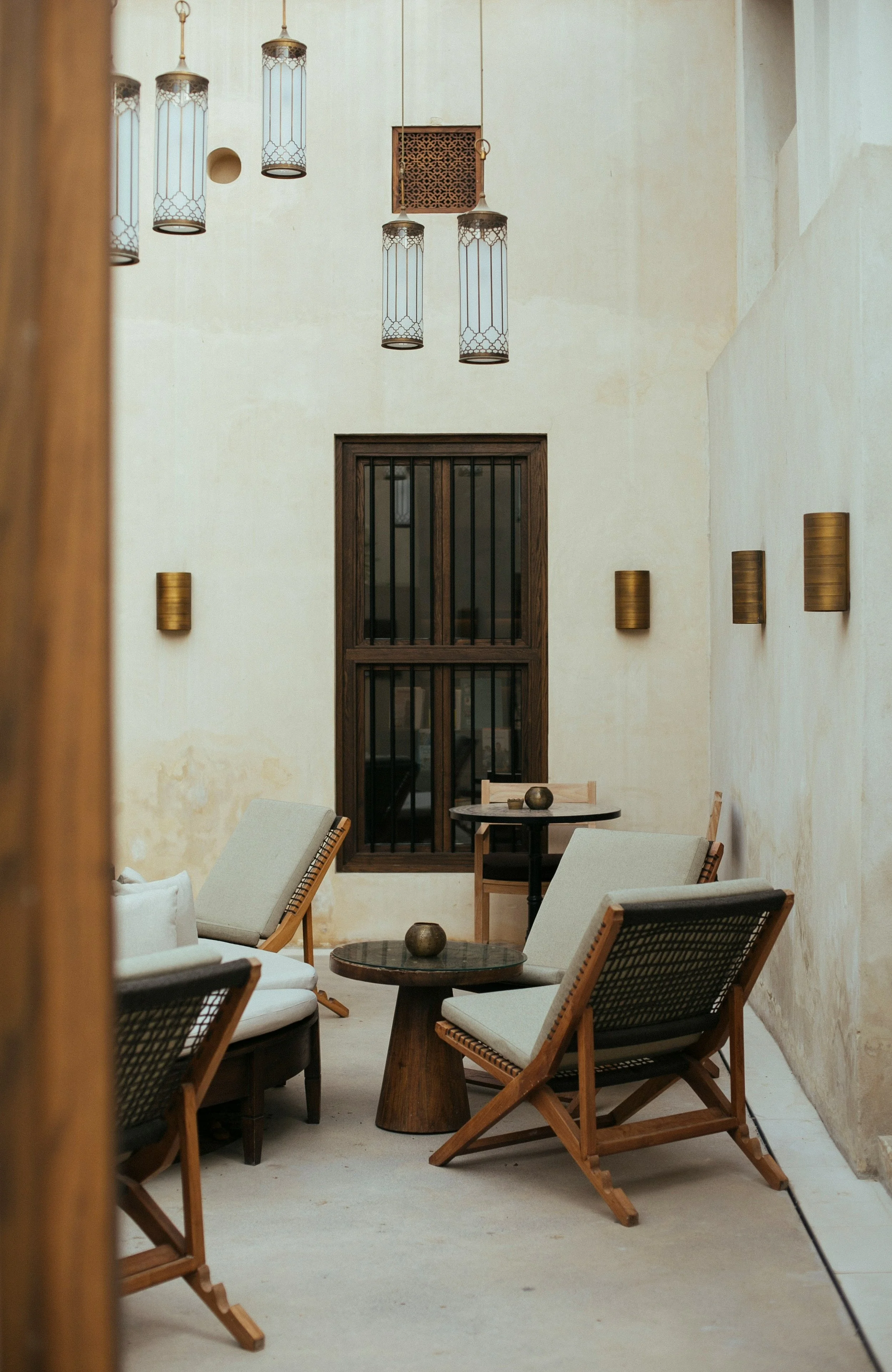 A cozy indoor seating area with wooden chairs, light-colored cushions, small round tables, decorative wall sconces, hanging lanterns, and a window with black bars.